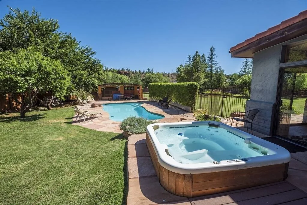 Backyard with swimming pool and hot tub, surrounded by green trees and lawn, under a clear blue sky.