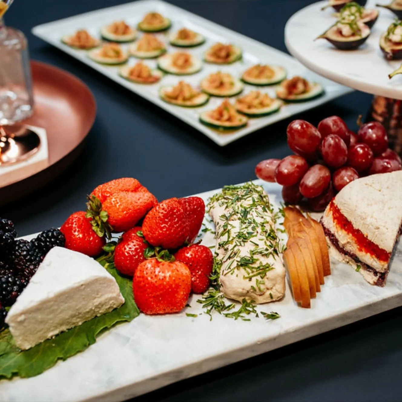 Assorted cheeses, strawberries, grapes, and crackers on a serving platter, with additional appetizers on plates in the background.