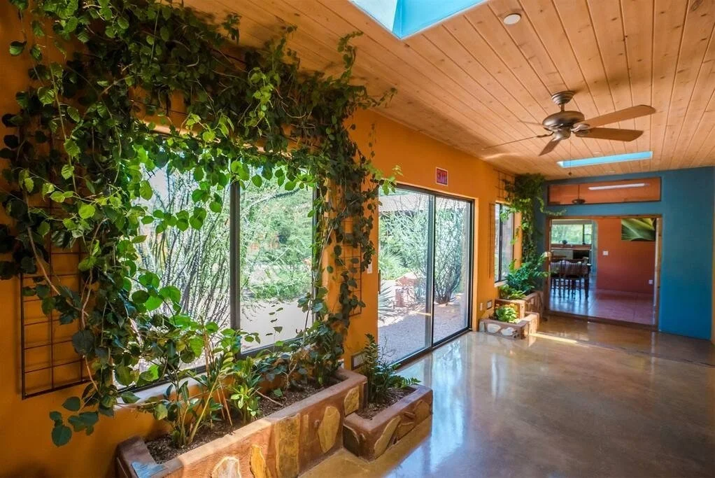 Bright living room with wood-paneled ceiling, large sliding glass doors, and vibrant yellow walls accented with green plants and garden views.