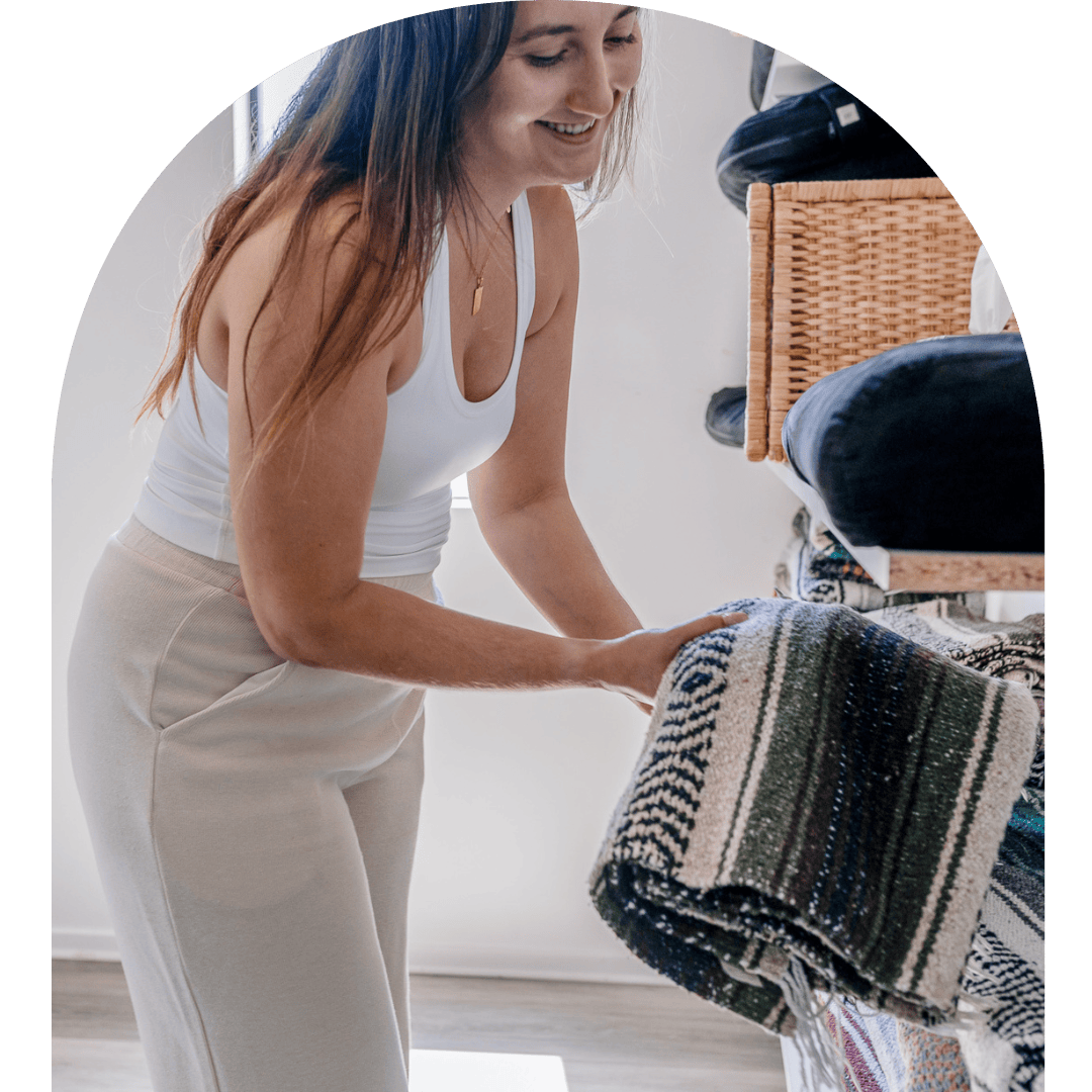 A woman with long brown hair wearing a white tank top and beige pants is folding a blanket in a room with shelves filled with folded clothes.