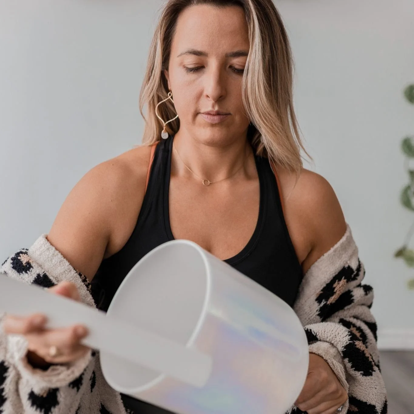 A woman with shoulder-length blonde hair wearing a black tank top and a beige and black patterned cardigan pouring a drink from a white pitcher.
