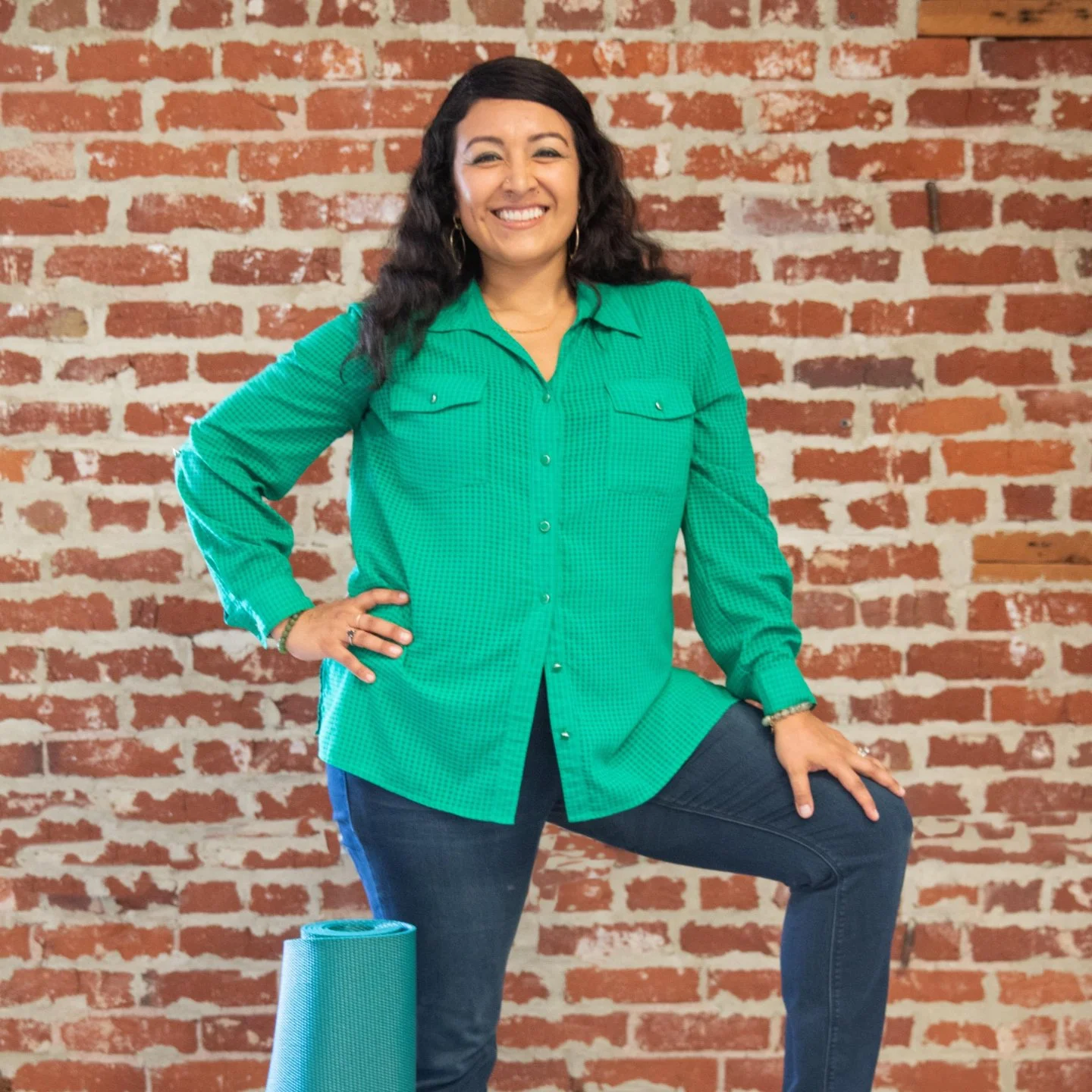 A woman with dark curly hair wearing a bright green long-sleeve shirt and blue jeans, standing against a red brick wall, smiling, with a blue yoga mat on the floor beside her.