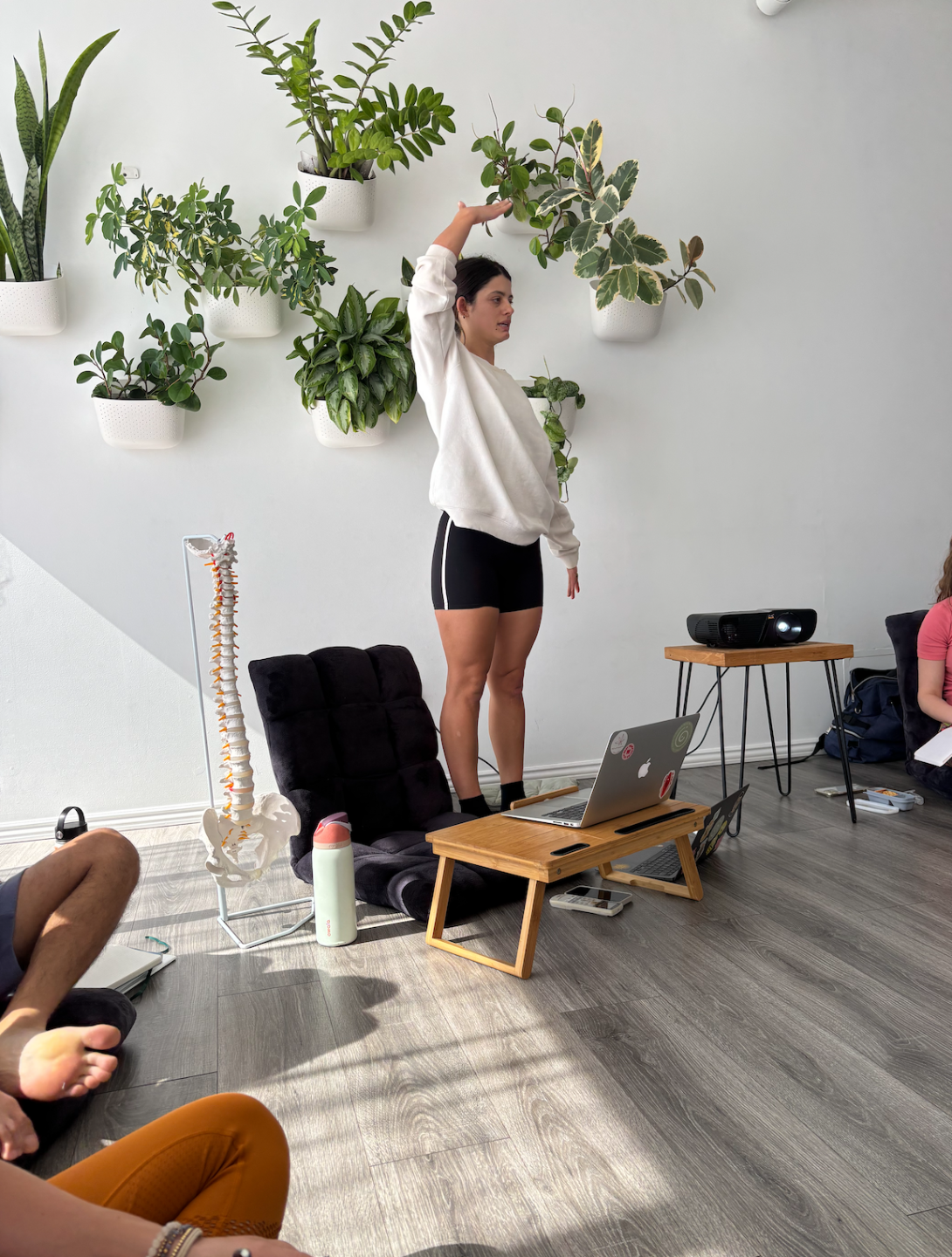 A woman is standing on a small wooden platform, stretching her arm overhead, in front of a white wall with hanging potted plants. There are people sitting on the floor, observing, and a model of a human spine is nearby. A laptop and a projector are on small tables.