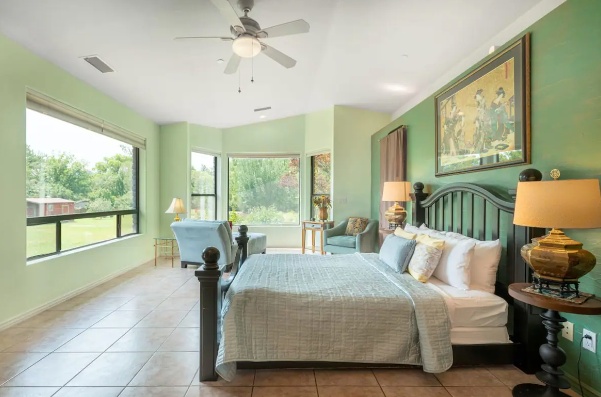Bright bedroom with large windows, a dark wooden bed with beige bedding, nightstands with lamps, a sitting area with two chairs and a side table, and a landscape painting on a green wall.