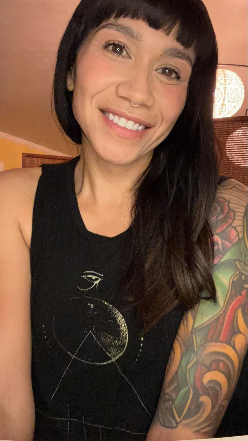 A smiling woman with dark hair and a septum piercing, wearing a black sleeveless shirt with a celestial design, showing a colorful tattoo on her arm, in an indoor setting with warm lighting.