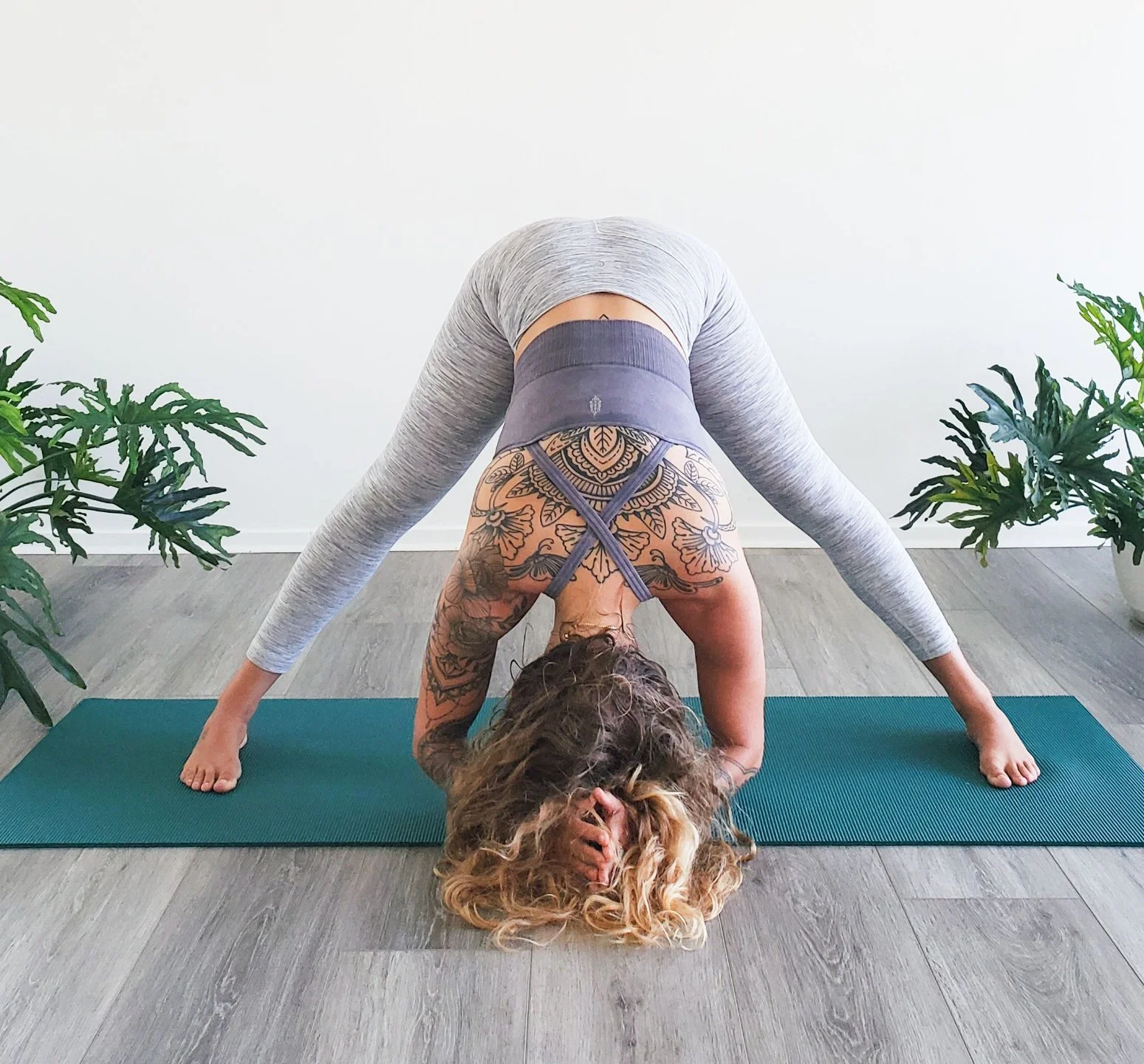 Woman practicing yoga in a forward bend pose with her head on the floor, on a yoga mat, surrounded by potted plants.