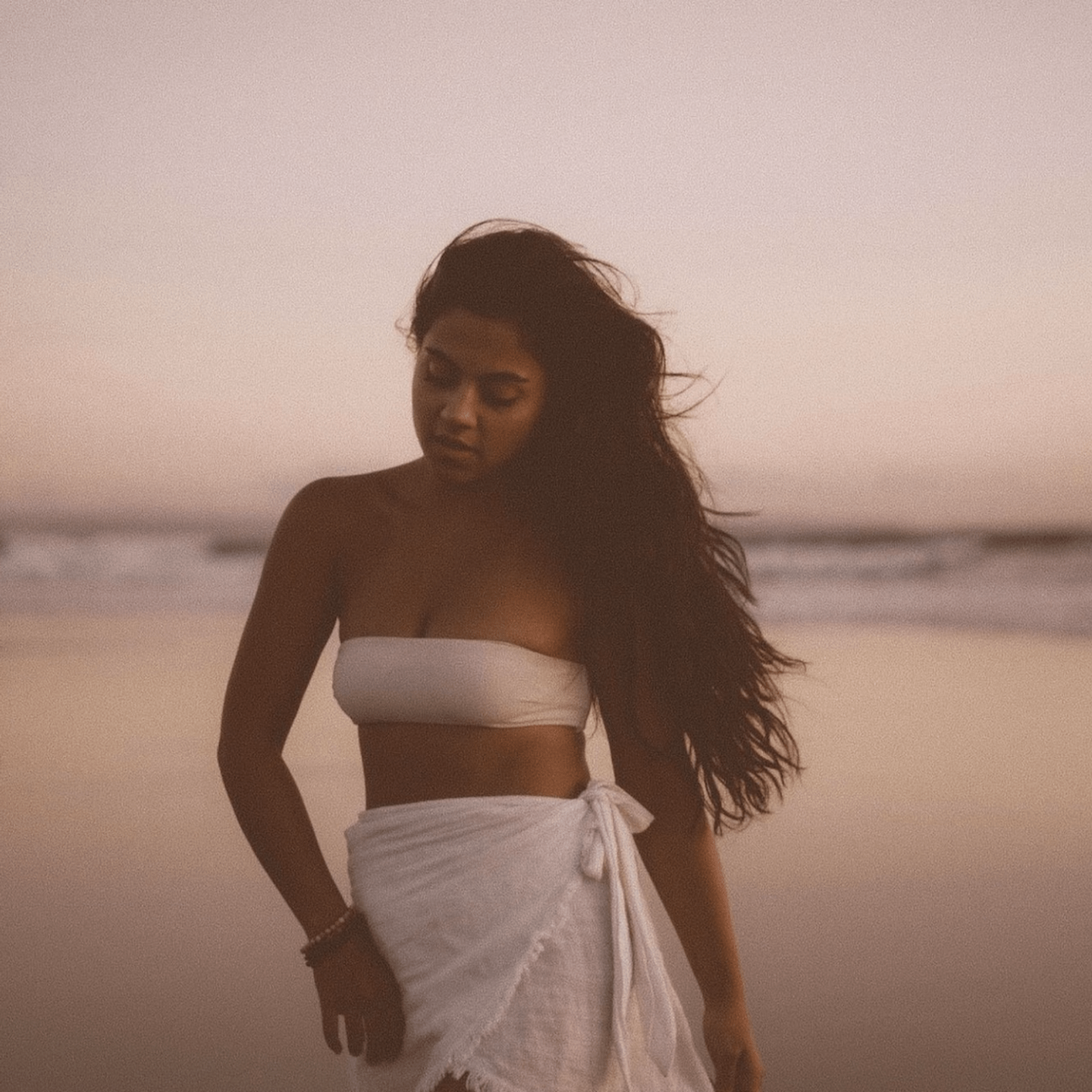 A woman stands on a beach during sunset, with her eyes closed and long hair blowing in the wind, wearing a white strapless top and a matching white wrap skirt.