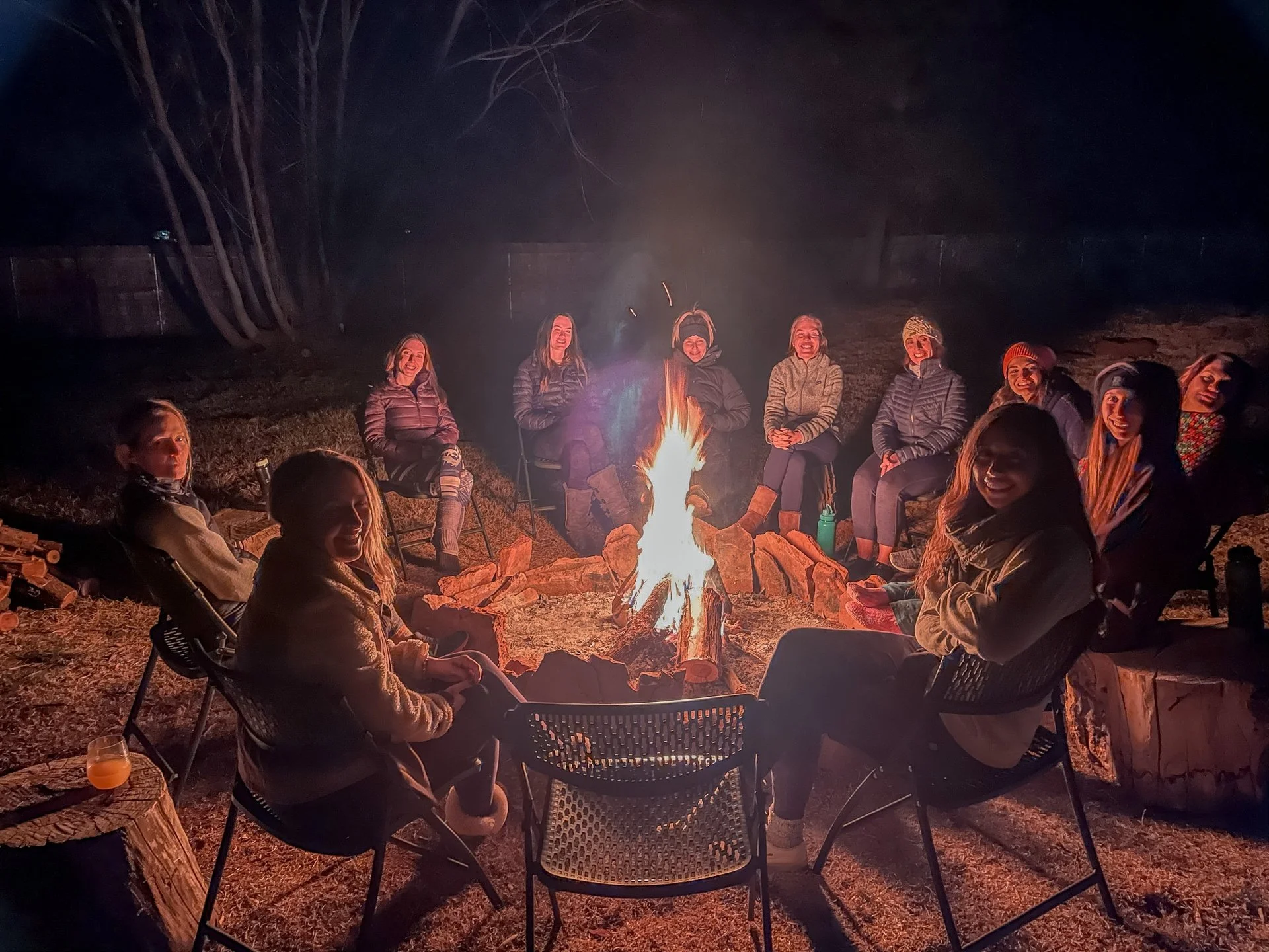 A group of people sitting around a campfire outdoors at night, smiling, with some holding drinks, in a circle.