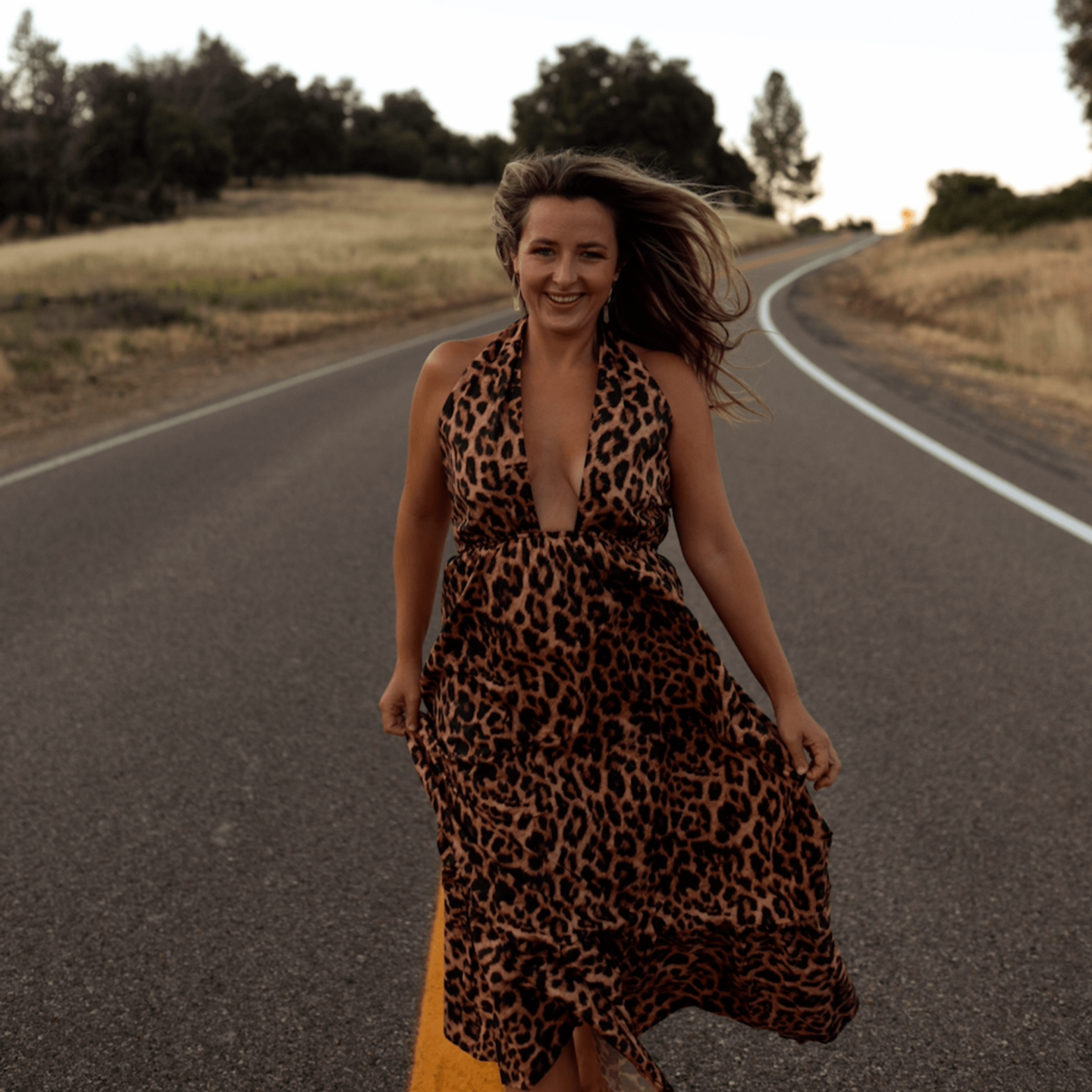 A woman with long hair, dressed in a leopard print dress, walking on a curved rural road during sunset, smiling and enjoying the moment.