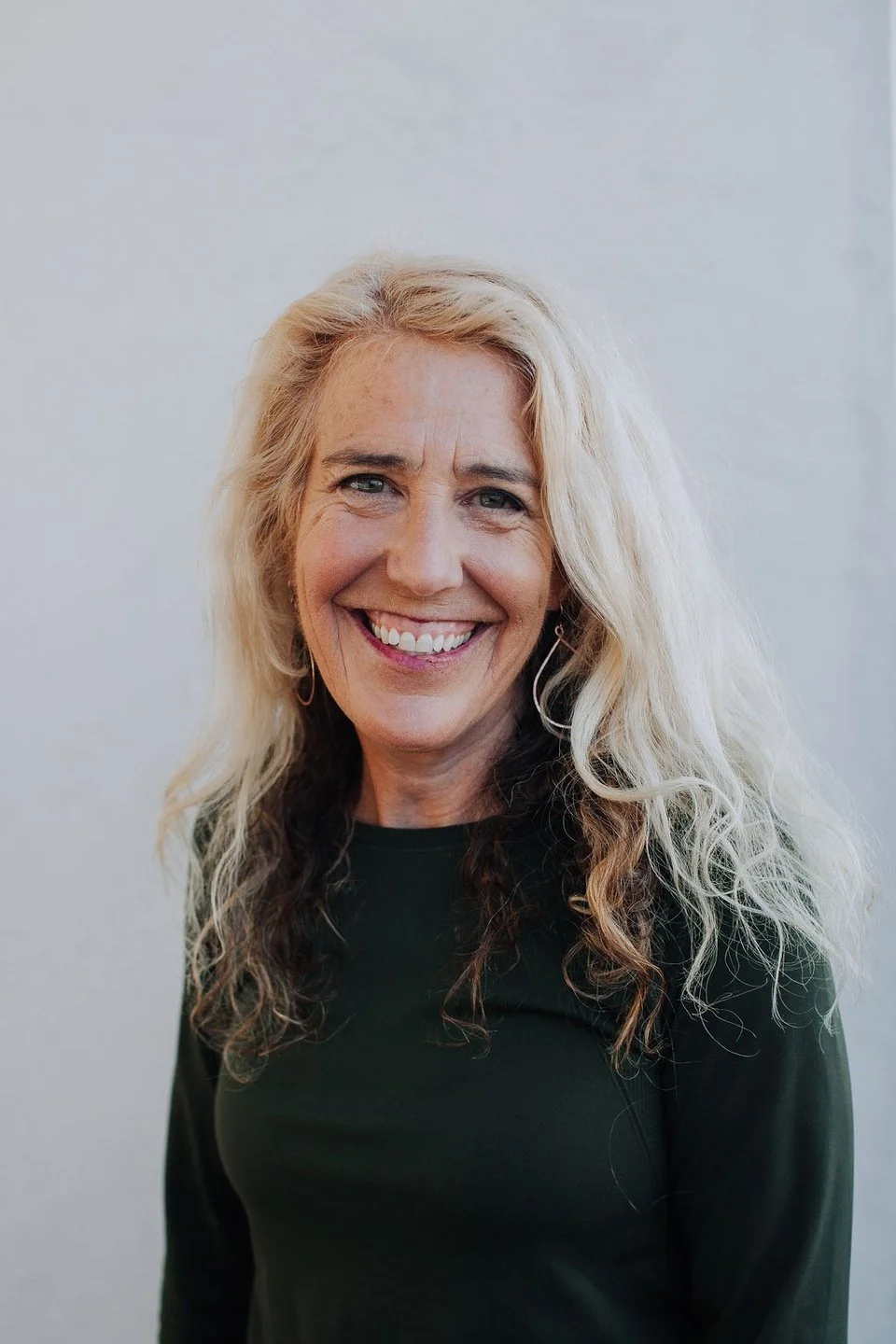 A smiling middle-aged woman with long, curly blonde hair, wearing a black top, standing against a light-colored background.