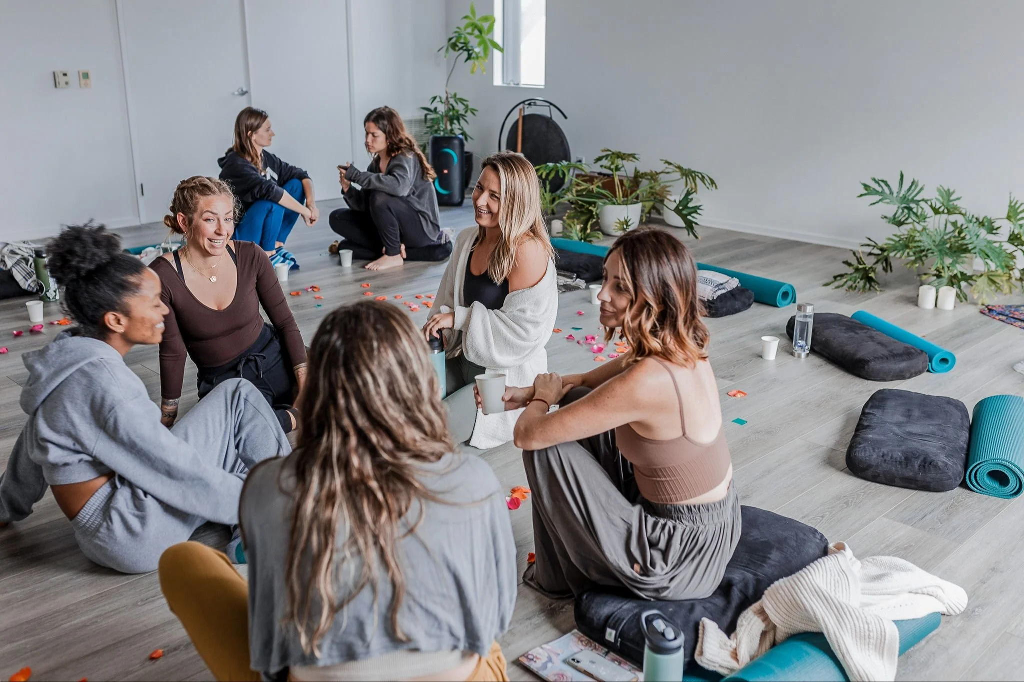 Group of women sitting on the floor, smiling and chatting in a yoga or wellness studio with mats and plants.