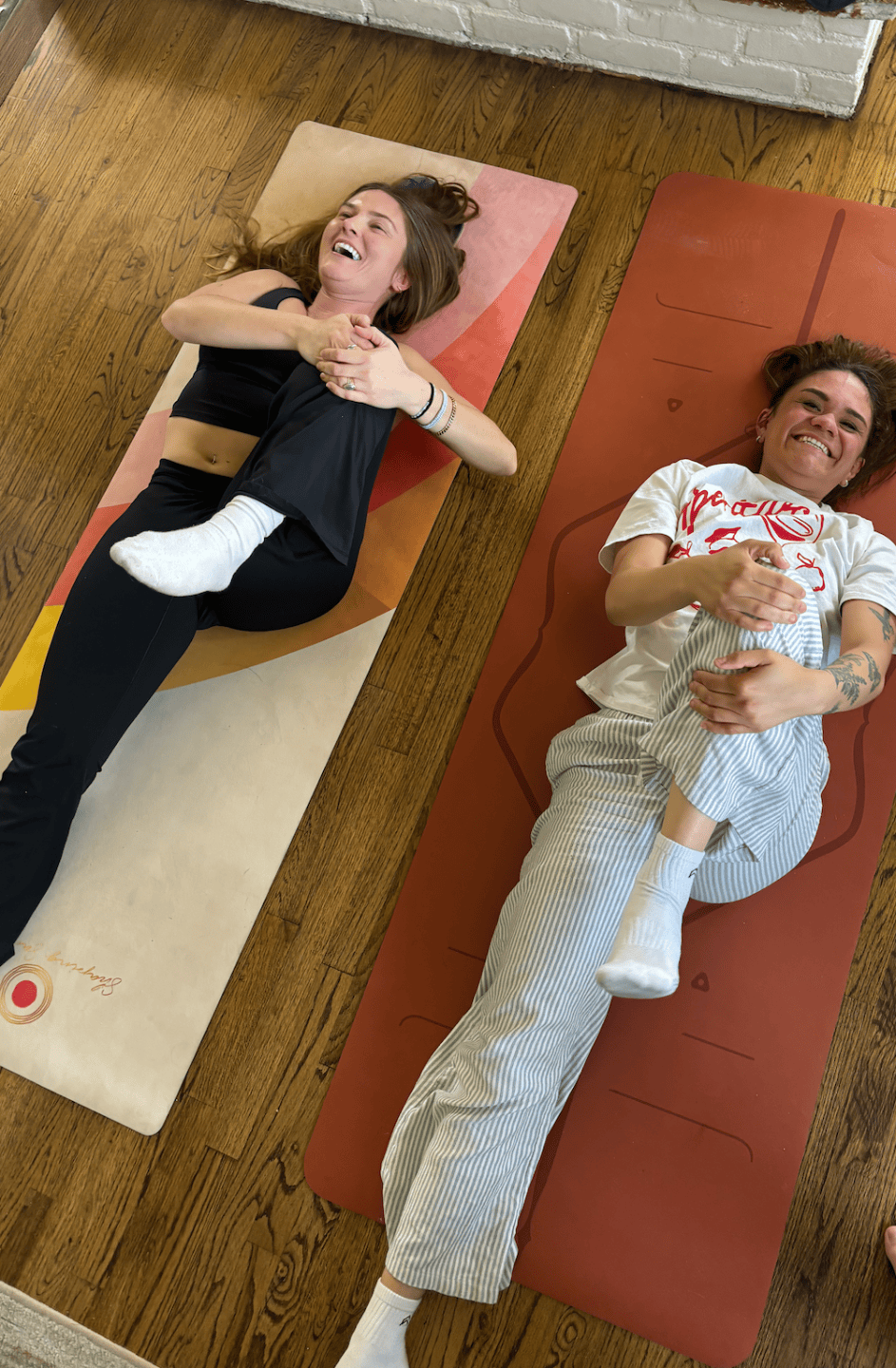 Two women lying on yoga mats on a wooden floor, laughing and playing with a baby, who is lying on the woman on the left.