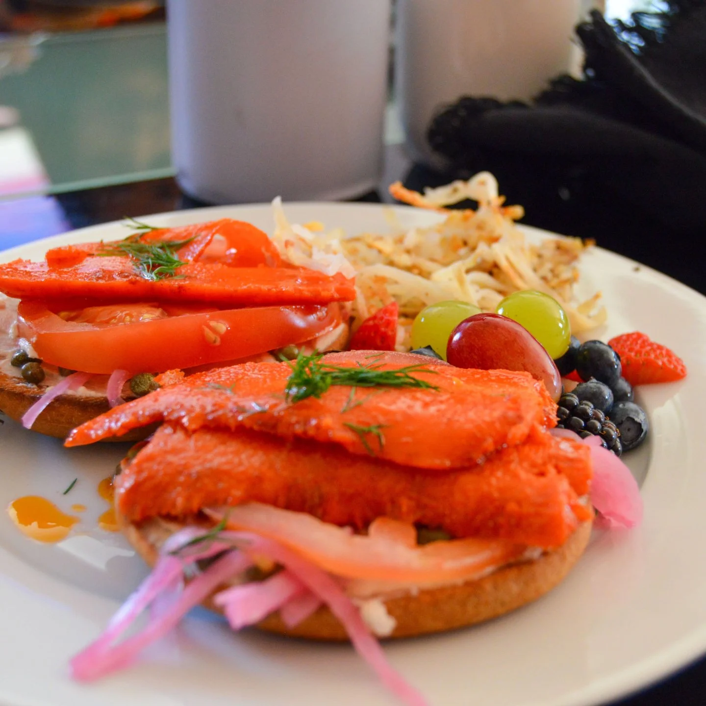 Plate with smoked salmon bagel, tomato, onion, and dill, along with a side of pasta salad and mixed fresh grapes, strawberries, and blueberries.