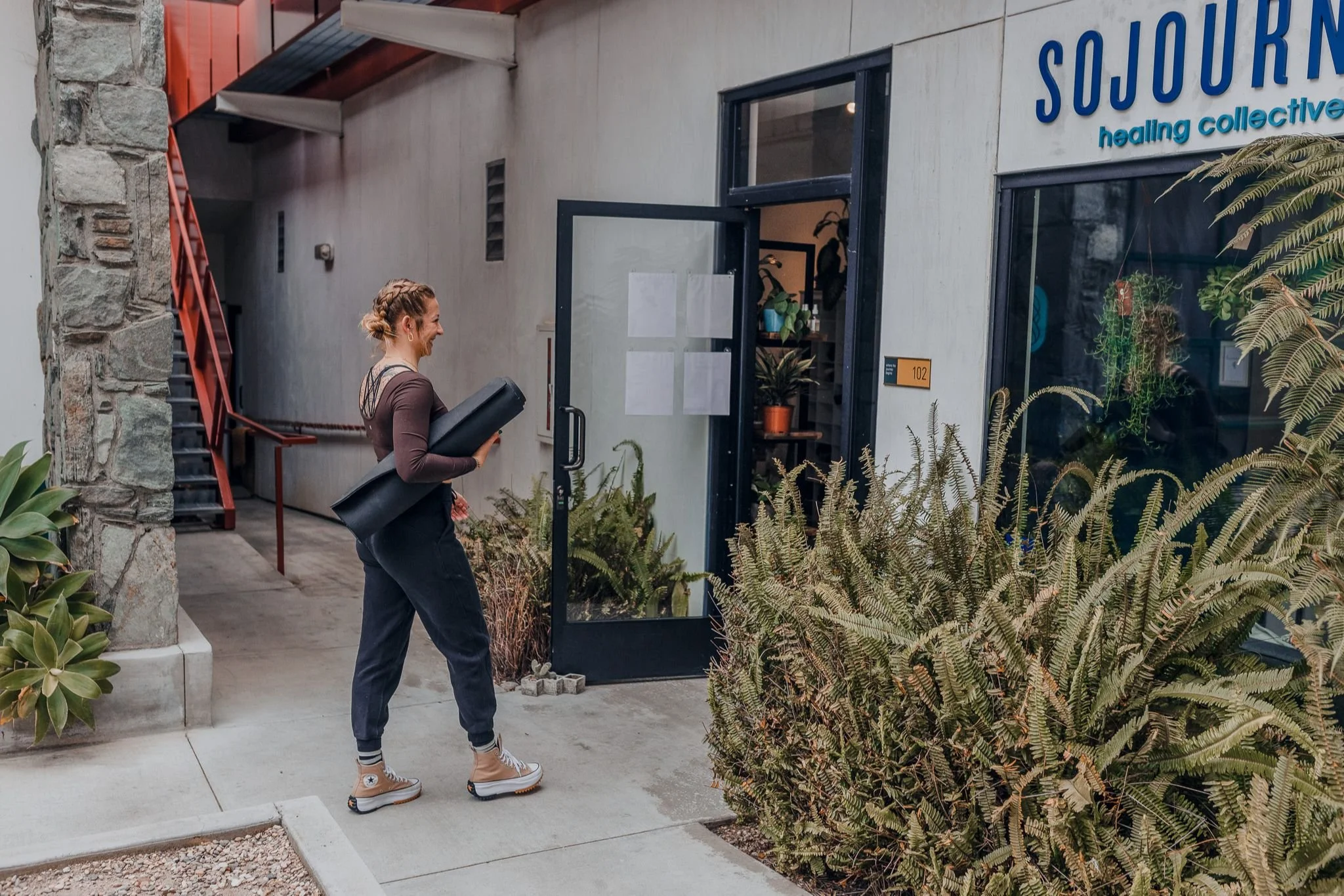 A woman with braided hair carrying a yoga mat walks into a wellness center named Sojourn Healing Collective.