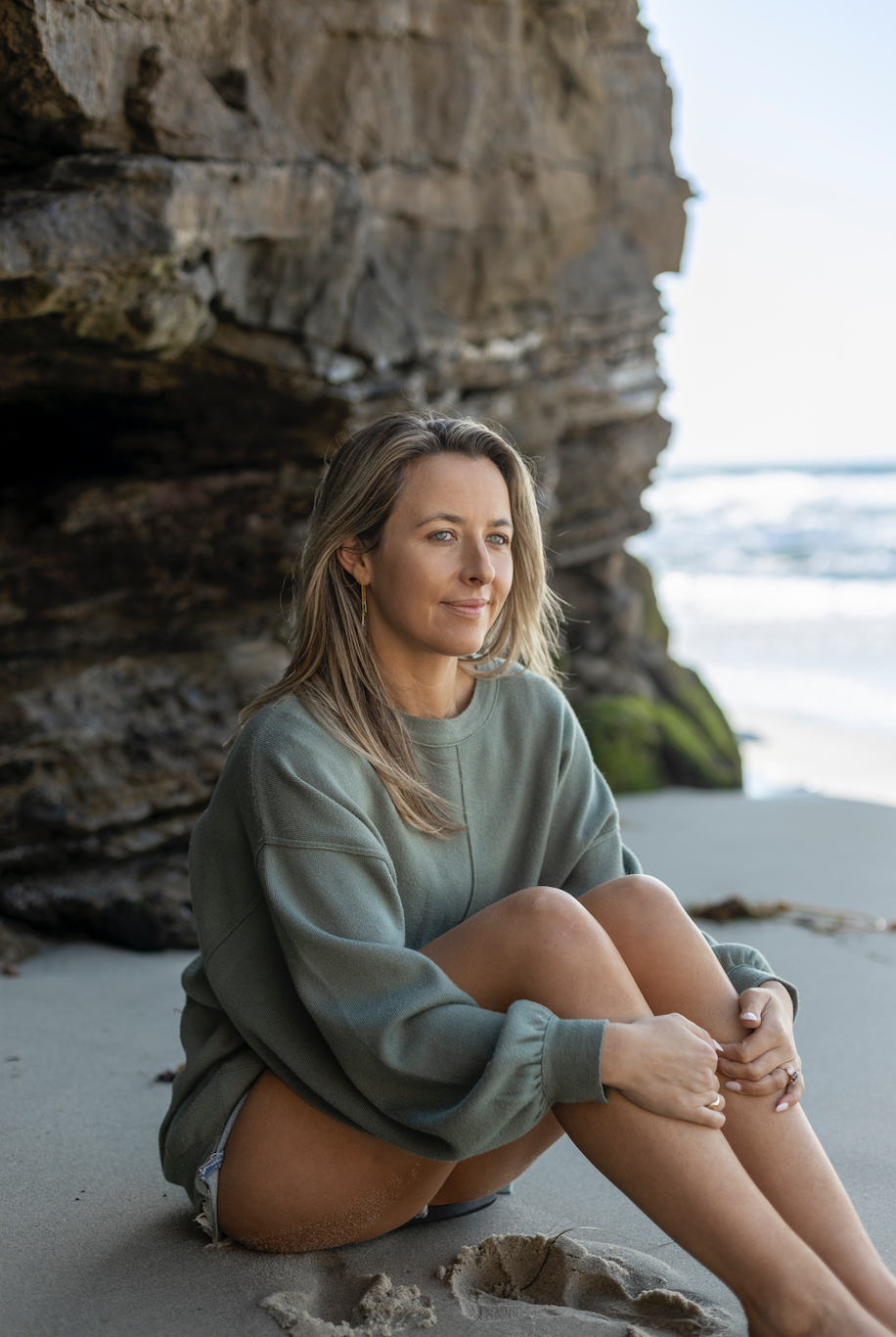 A woman sitting cross-legged on the beach near a rocky cliff, with ocean waves in the background.