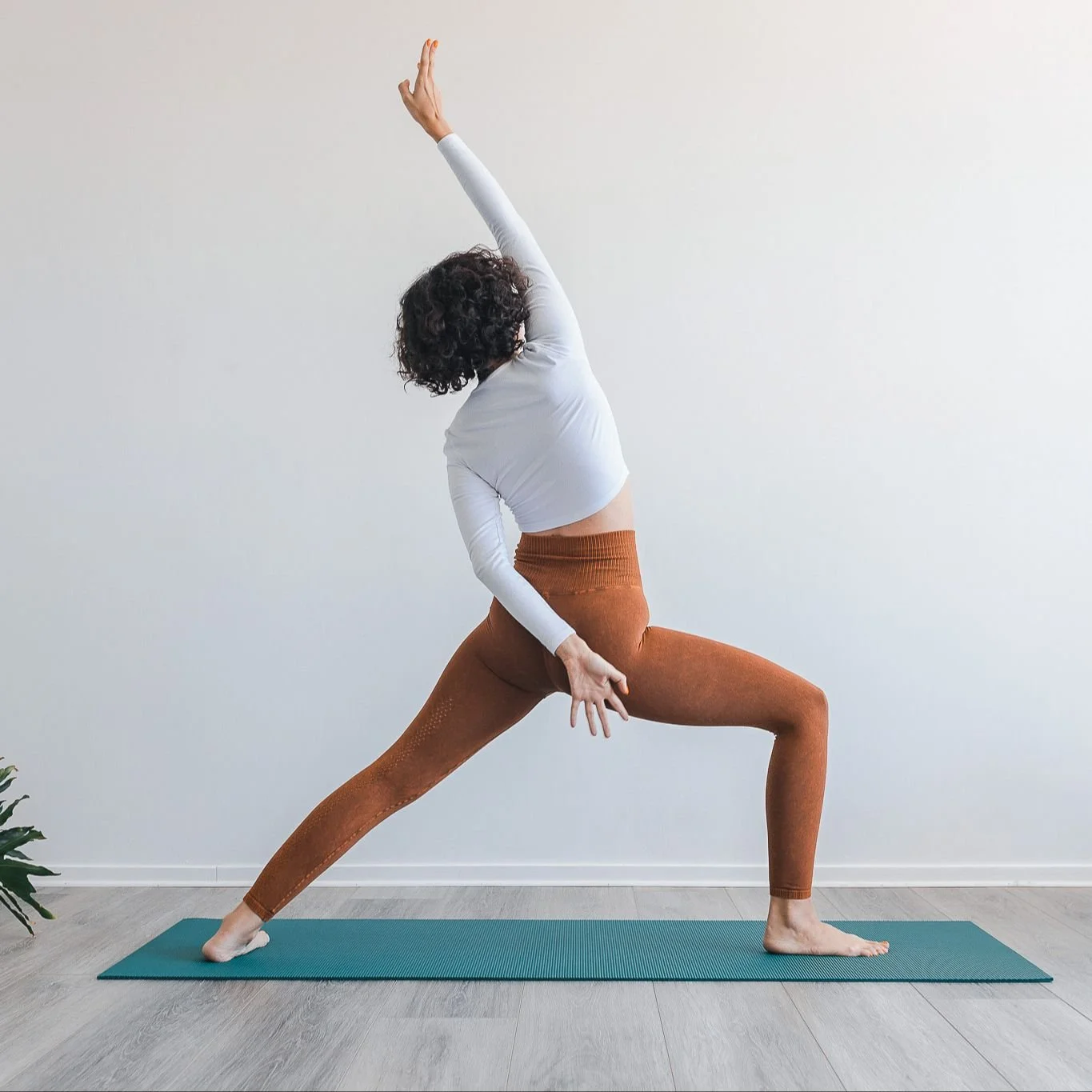 A woman practicing yoga, in a lunge pose with one arm reaching upward and the other hand resting on her thigh, on a yoga mat indoors.
