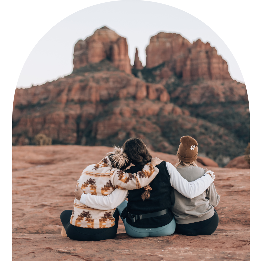 Three friends sitting on a rocky desert landscape with red rock formations in the background, embracing each other and watching the scenery.