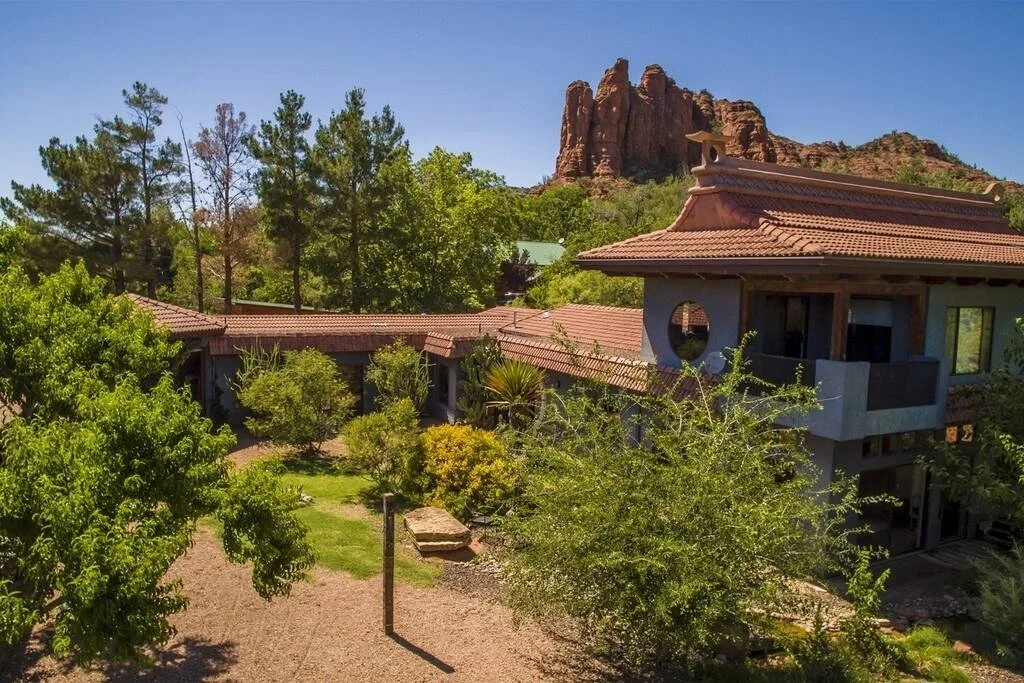 A house with a red tile roof and balconies surrounded by trees and shrubs, with red rock formations in the background under a clear blue sky.