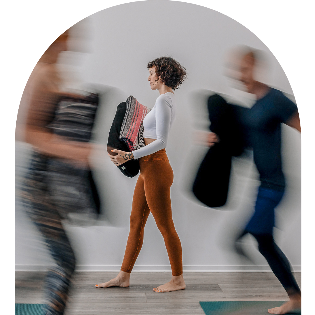 A woman in white and brown leggings walking barefoot while holding a folded blanket, with two blurred people walking past her in a hallway.
