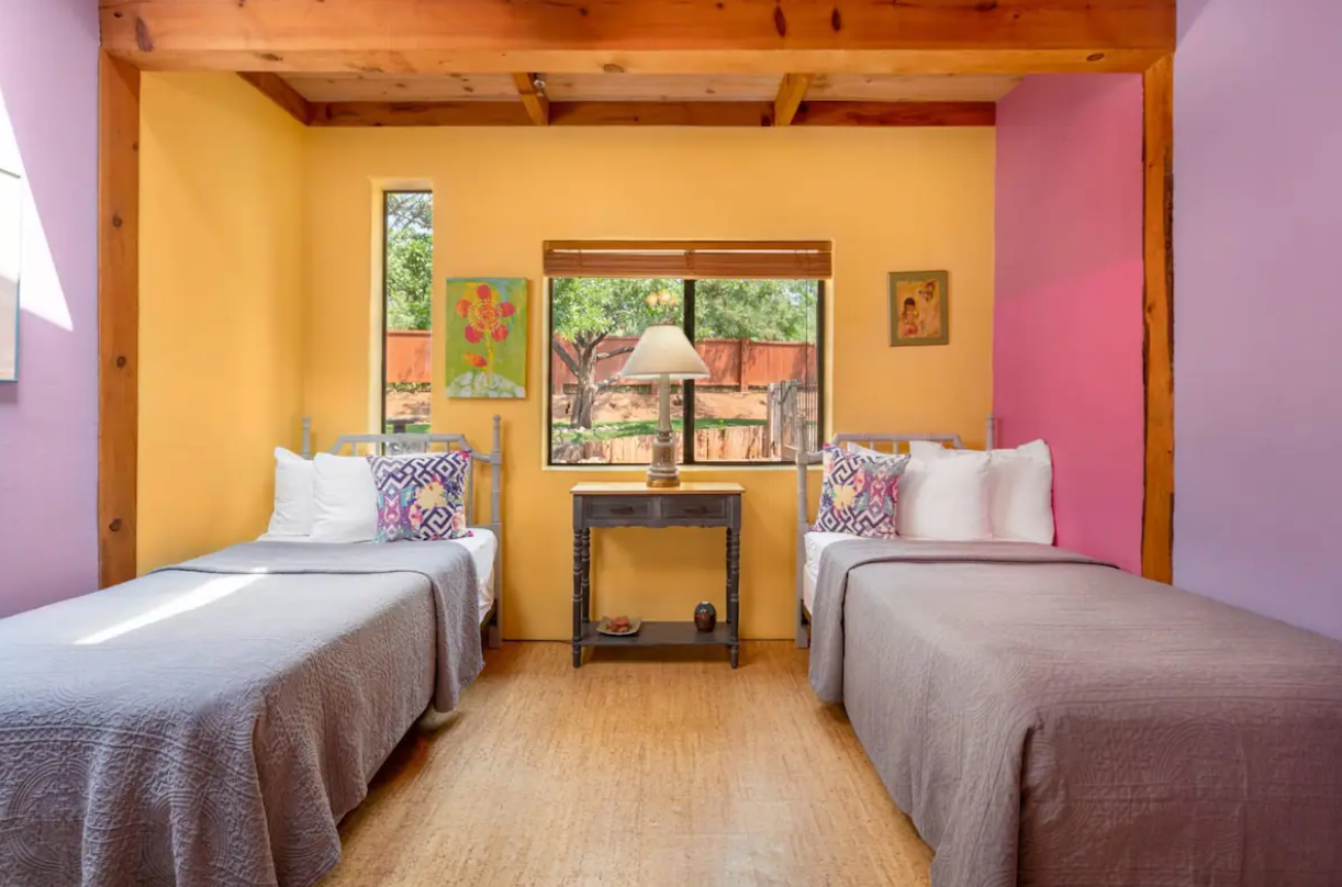 A bedroom with two beds, a window with a view of trees, colorful wall paintings, a lamp on a small table, and wood beam ceiling.