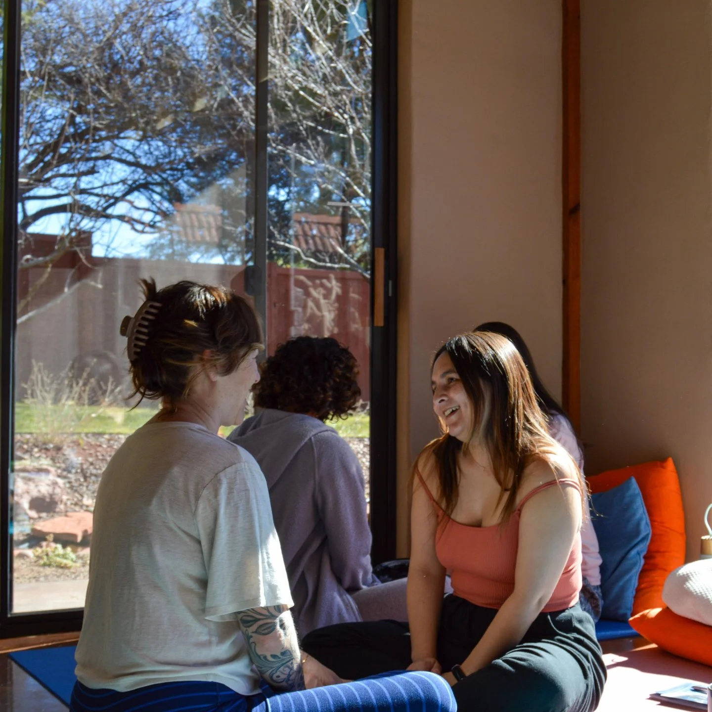 Group of women sitting and chatting indoors near a large window with a garden view.