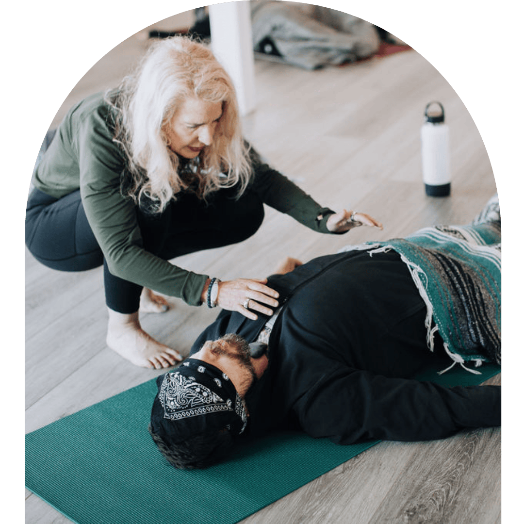 A woman with long blonde hair performing CPR on a man lying on a green exercise mat, with a water bottle nearby in a room with wooden flooring.