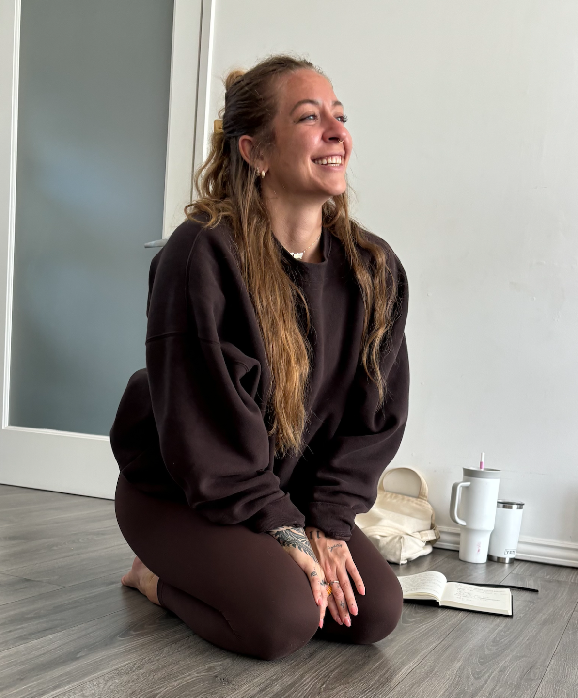 A woman with long, wavy hair, tattoos on her hands, and a nose ring, smiling while kneeling on a wooden floor. She wears a dark oversized sweatshirt and leggings. In the background, there are reusable drink containers, a bag, and an open notebook.