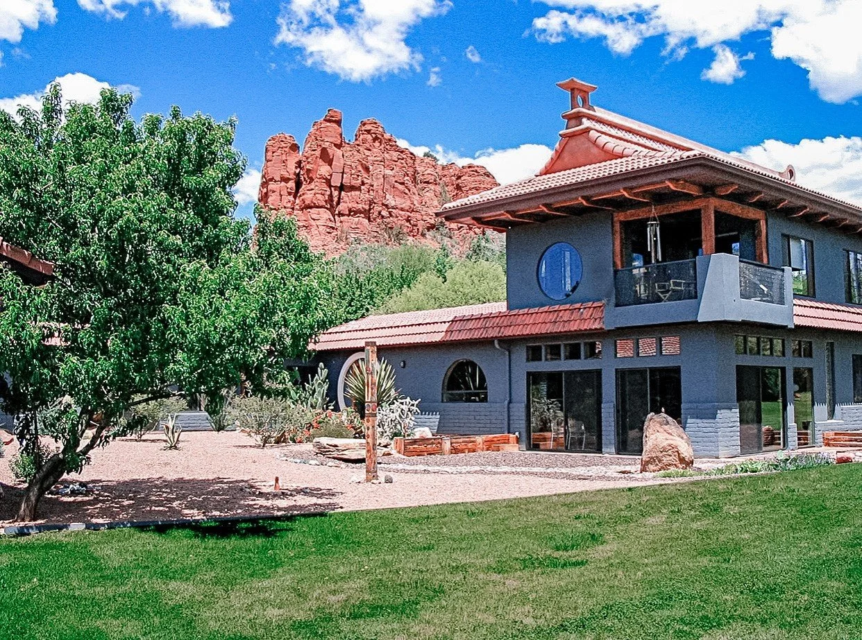 A modern house with a red tile roof, blue walls, and large windows, surrounded by green grass and trees, with red rock formations in the background under a partly cloudy sky.