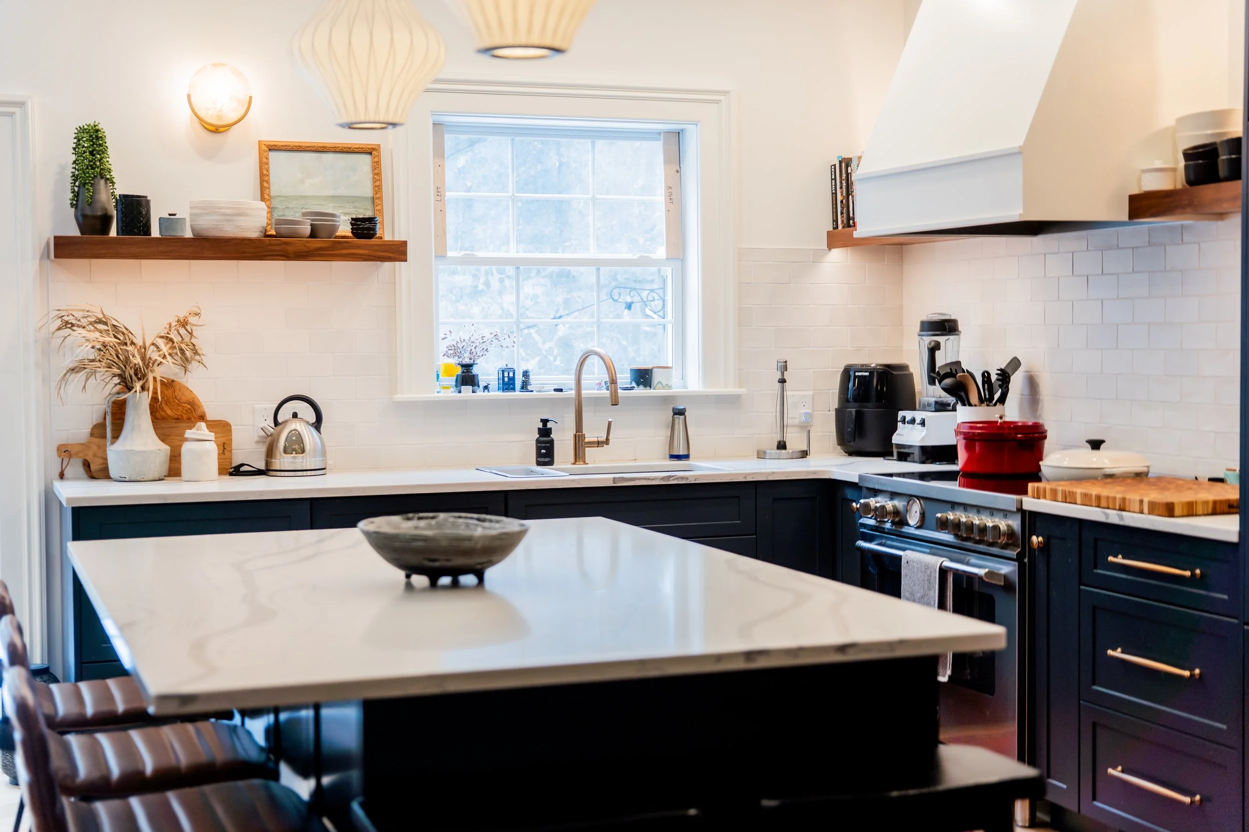 A modern kitchen with navy blue cabinets, white countertops, and wall tiles. There is a window above the sink, with various small decorative items on the windowsill. Open shelves hold bowls, books, and potted plants. The kitchen island has a ceramic bowl on top, and a dining table with chairs is visible in the foreground. Appliances and kitchen tools are on the counters, including a kettle, air fryer, and blender.