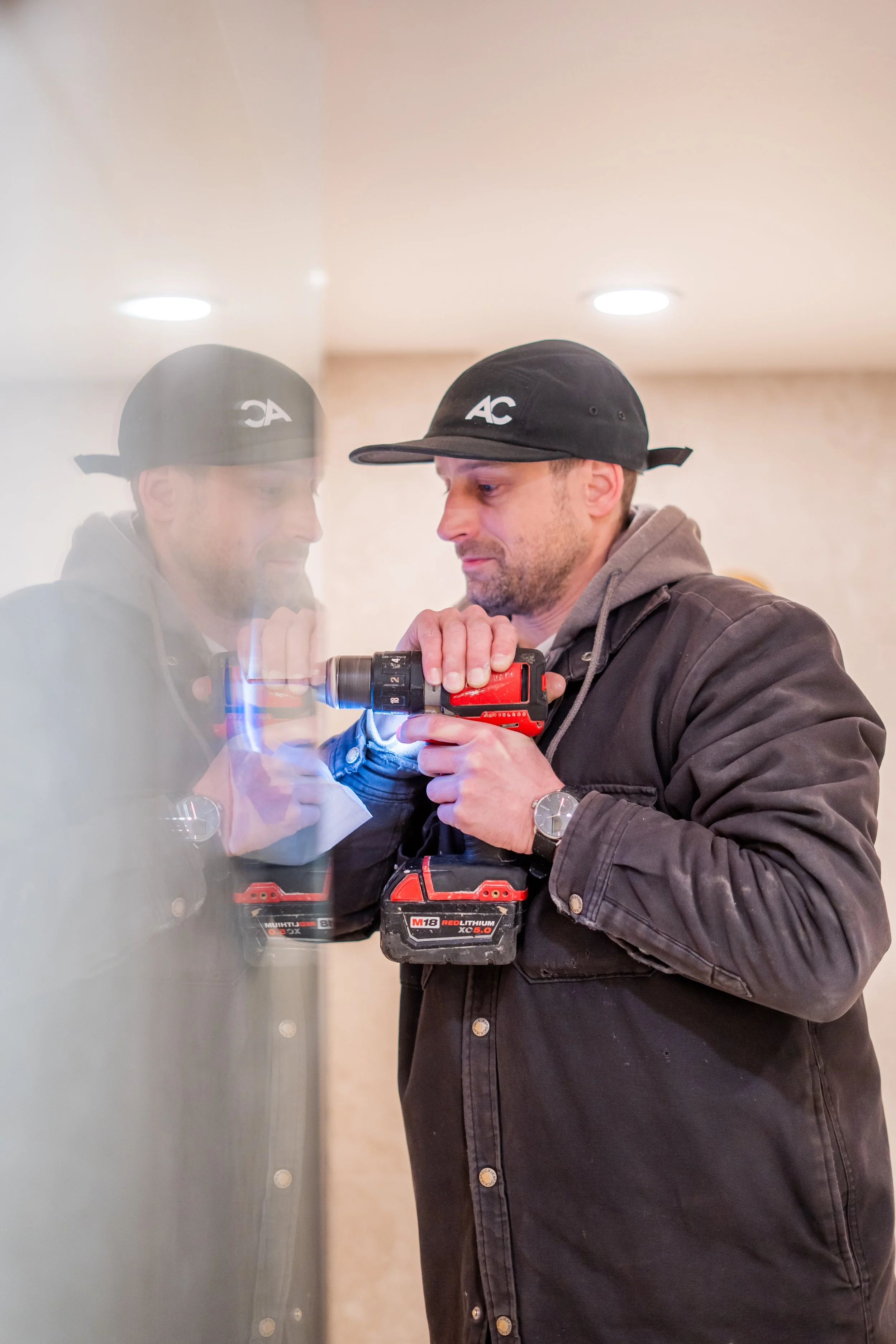 A man using a cordless power drill to fix or install something, with his reflection visible on a glass surface.