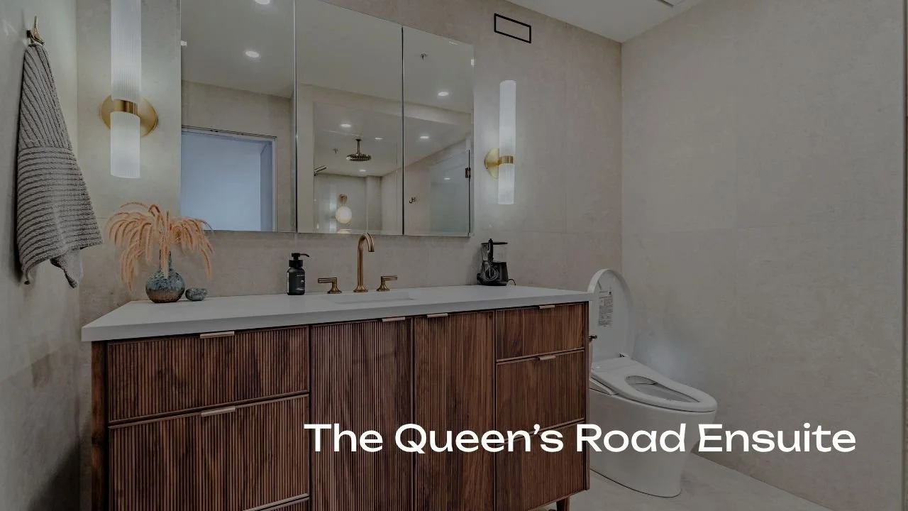 Modern bathroom with beige walls, a large mirror, a wooden vanity with a white countertop, a gold faucet, a wall-mounted light fixture, a decorative vase with dried plants, a soap dispenser, a hairdryer, and a high-tech toilet, with the text 'The Queen's Road Ensuite'