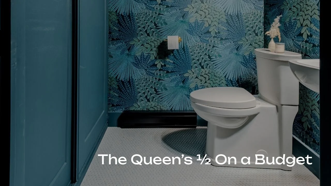 A cozy bathroom with blue tropical leaf-patterned wallpaper, white toilet, teal cabinetry, and a small side table with a white vase and decorative items.