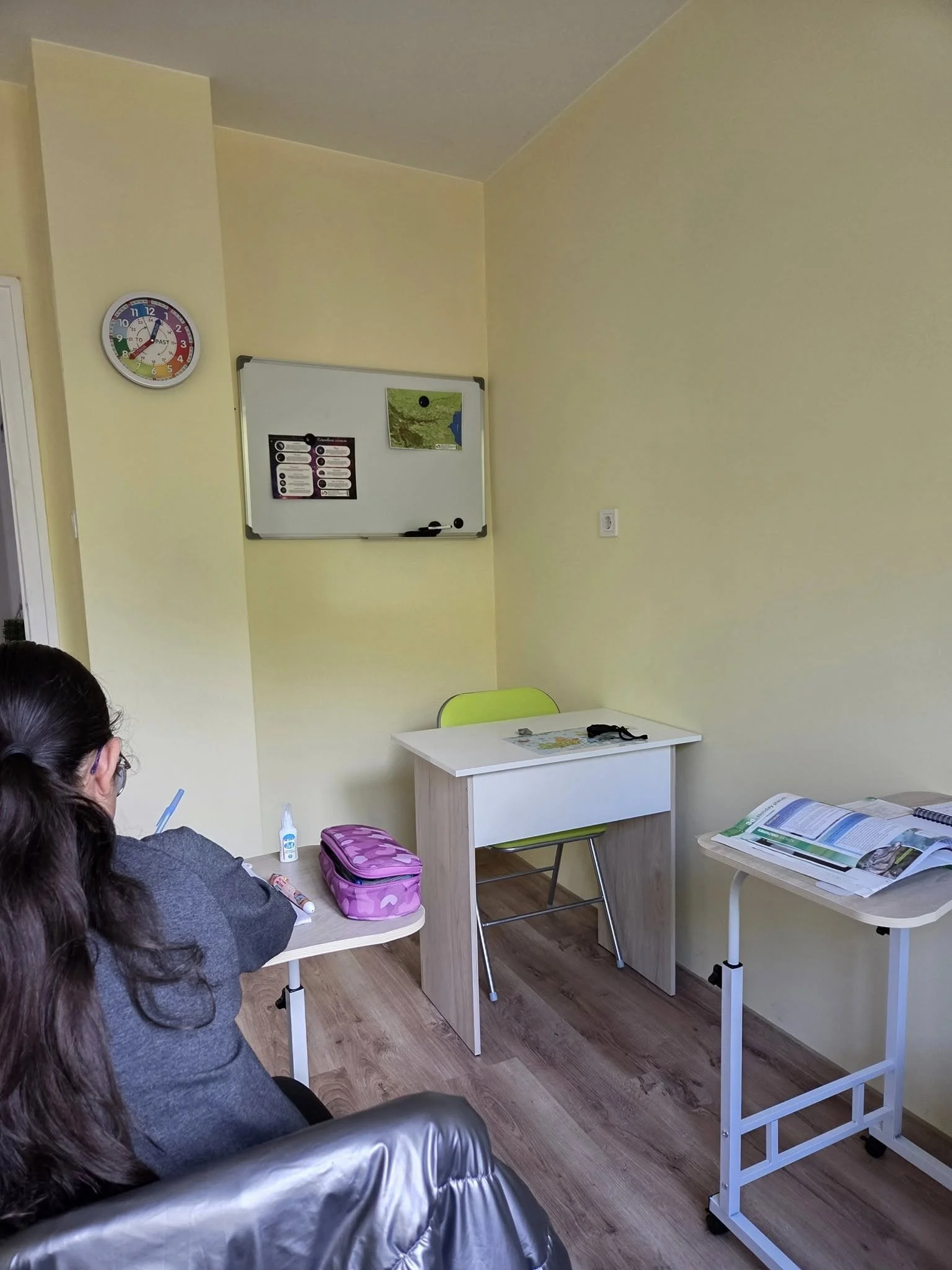 Empty classroom with a small desk, a partner desk, and a table with textbooks and notebooks. A person with long dark hair and glasses is seated on the left side of the image. A whiteboard with a map is mounted on the yellow wall, and a round multicol