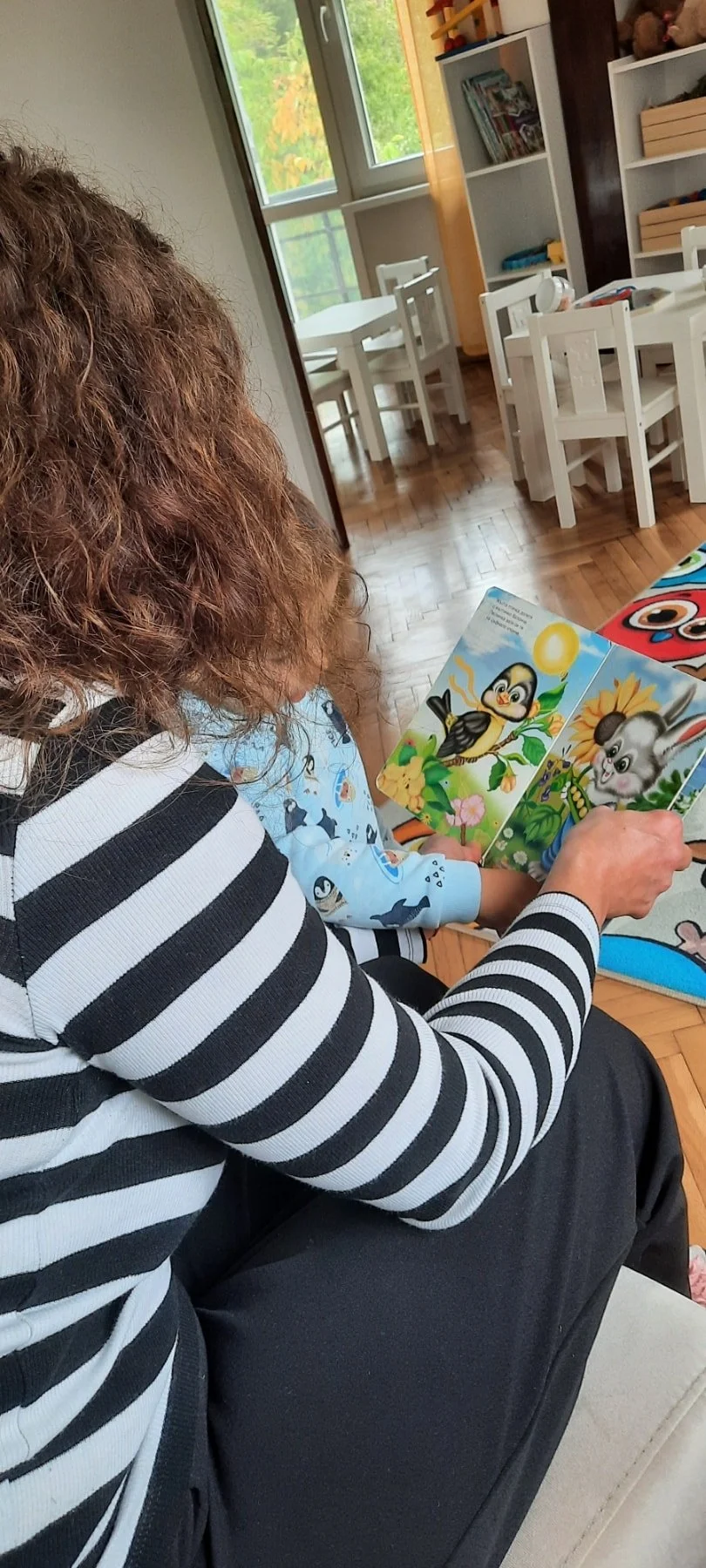 A woman with curly hair wearing a black and white striped shirt reading a children's picture book with a child in a blue penguin-themed pajamas, sitting on a beige sofa in a room with wooden floors and white furniture.