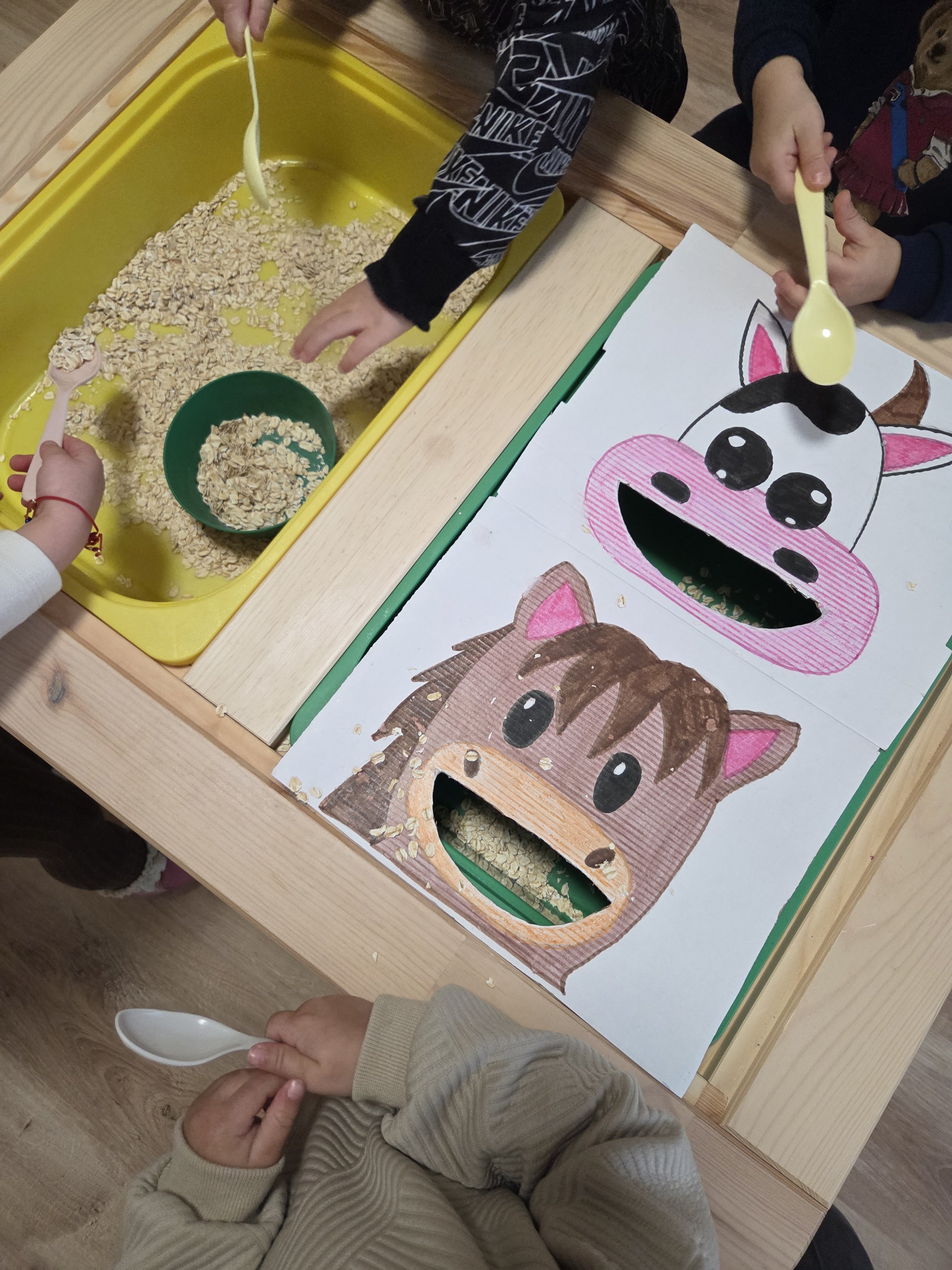 Children playing a game resembling a cow and horse with cereal or oatmeal through cutouts in paper faces on a table.