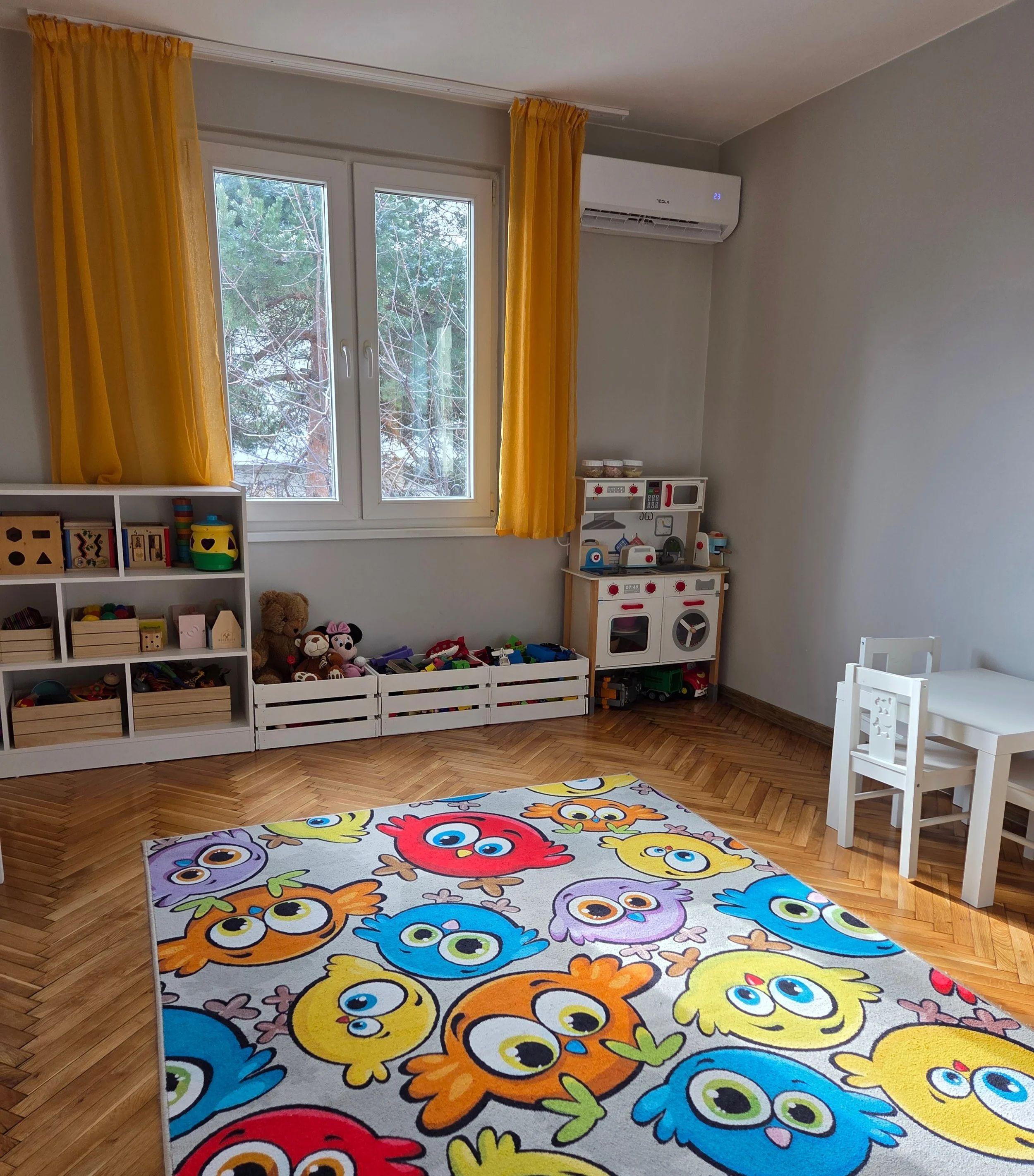 Colorful children's playroom with a owl-themed rug, white furniture, yellow curtains, and toy storage against the wall.