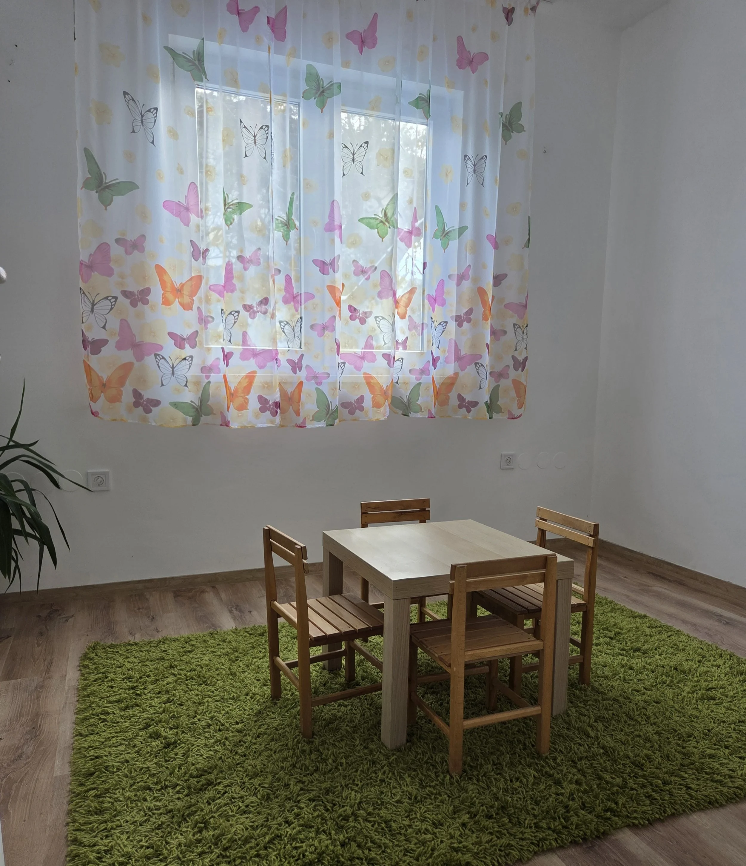 Small wooden table with four matching chairs on a green shag rug in a bright room with butterfly patterned curtains.