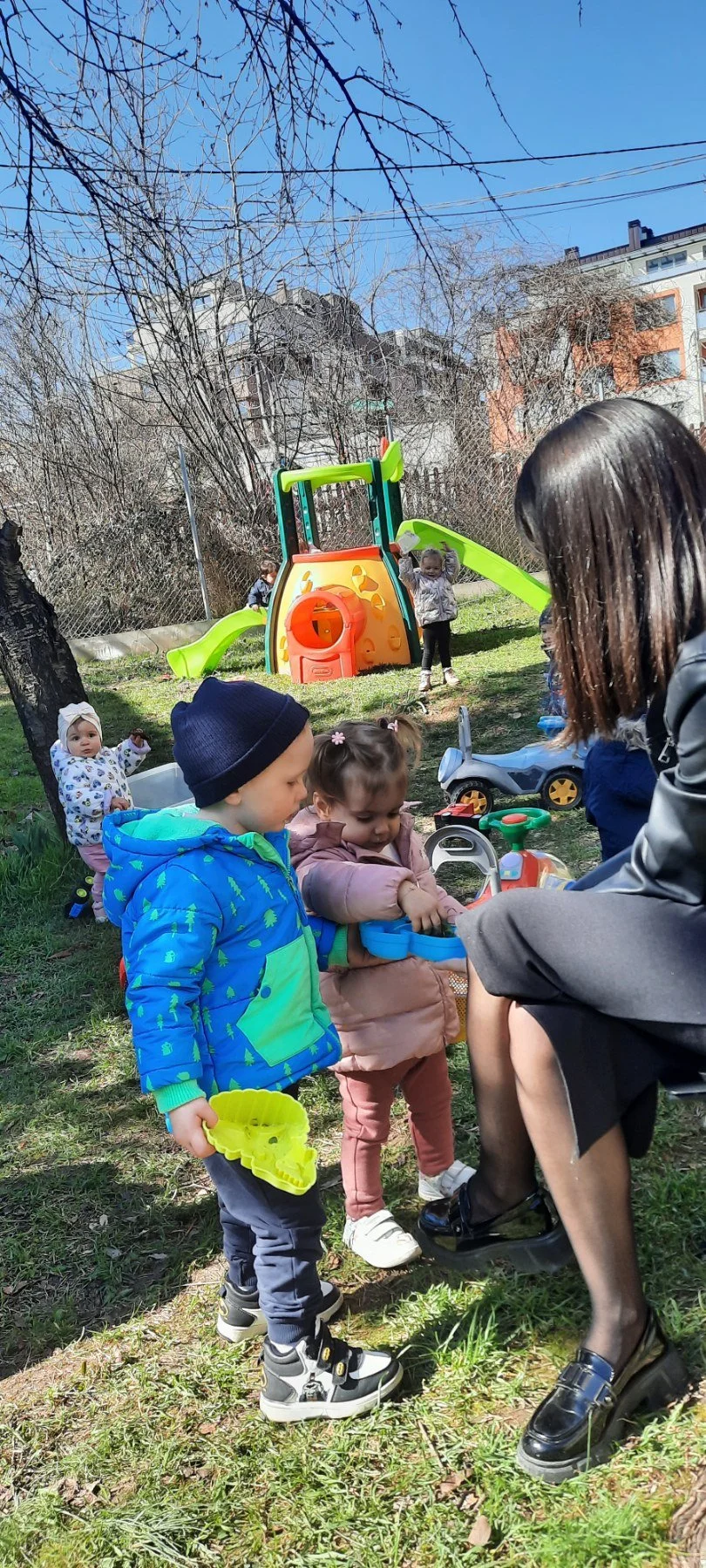 Children playing outside on a grassy area near a colorful playground with slides and climbing structures, supervised by an adult woman.