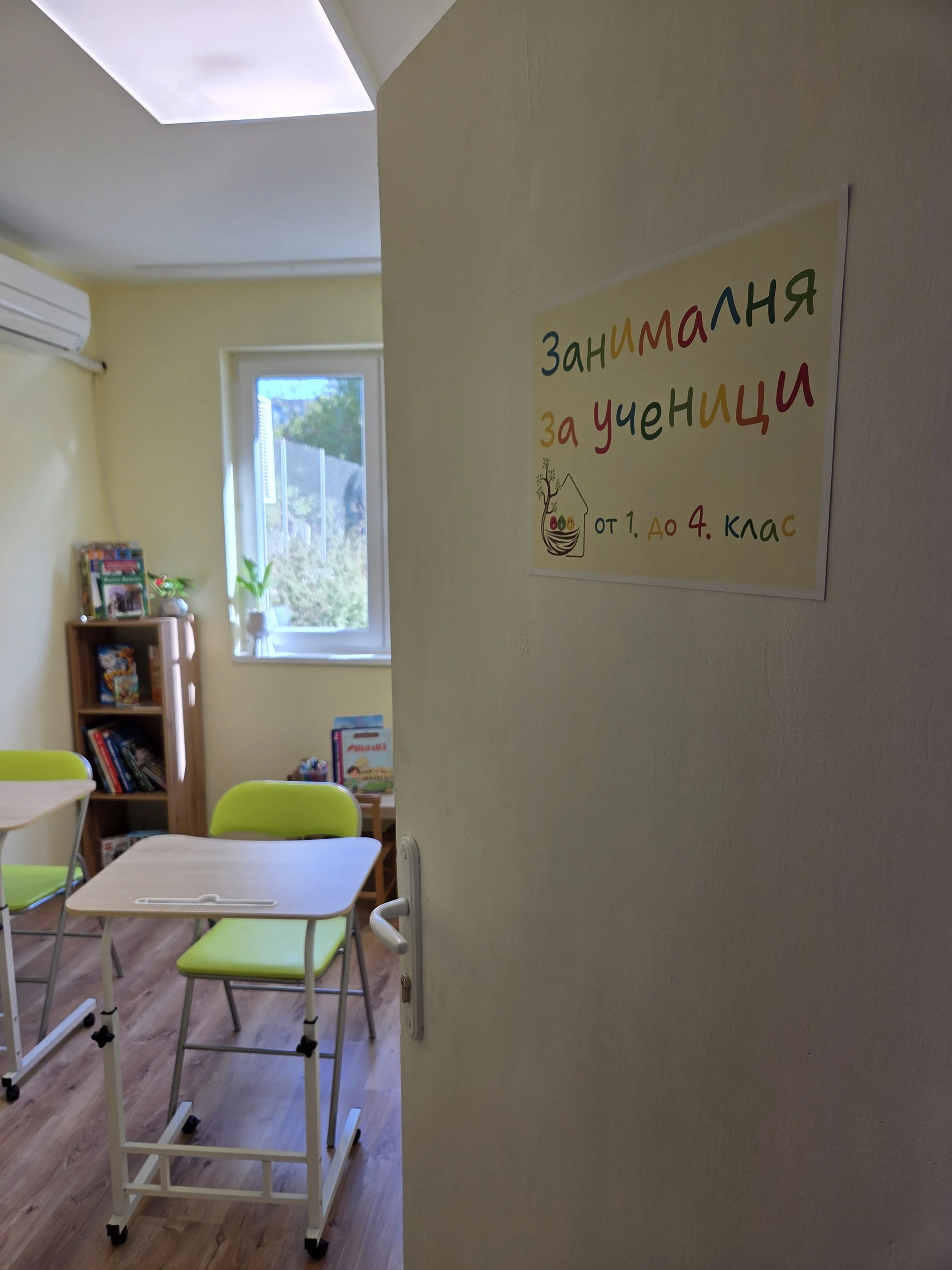 View of a classroom seen through a doorway with light yellow walls, a window showing trees outside, green desks and chairs, and shelves with books and decorations. A colorful sign on the door indicates the classroom is for students from 1st to 4th gr