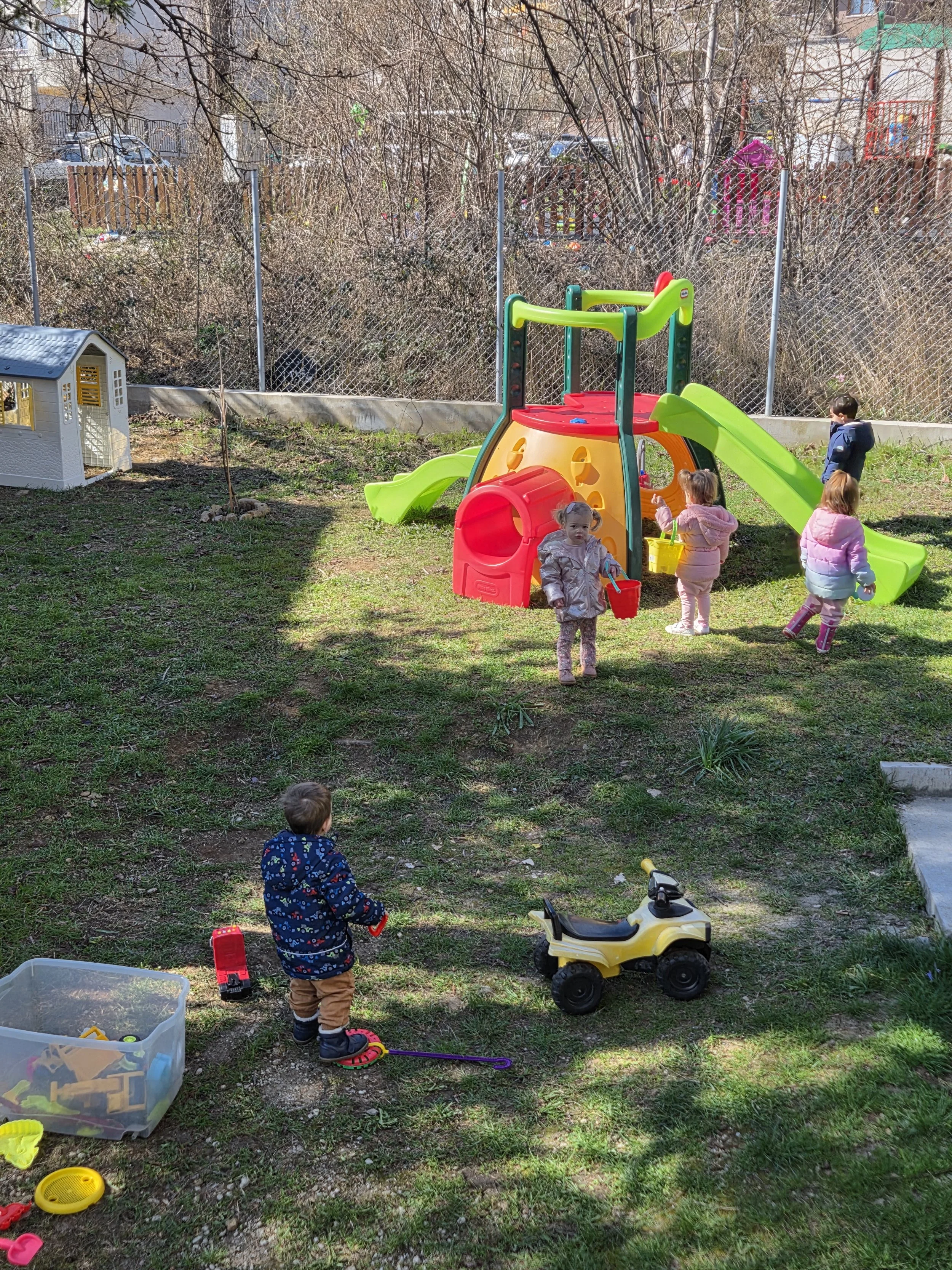 Children playing in an outdoor playground with a small slide and sandbox, surrounded by a chain-link fence and trees.