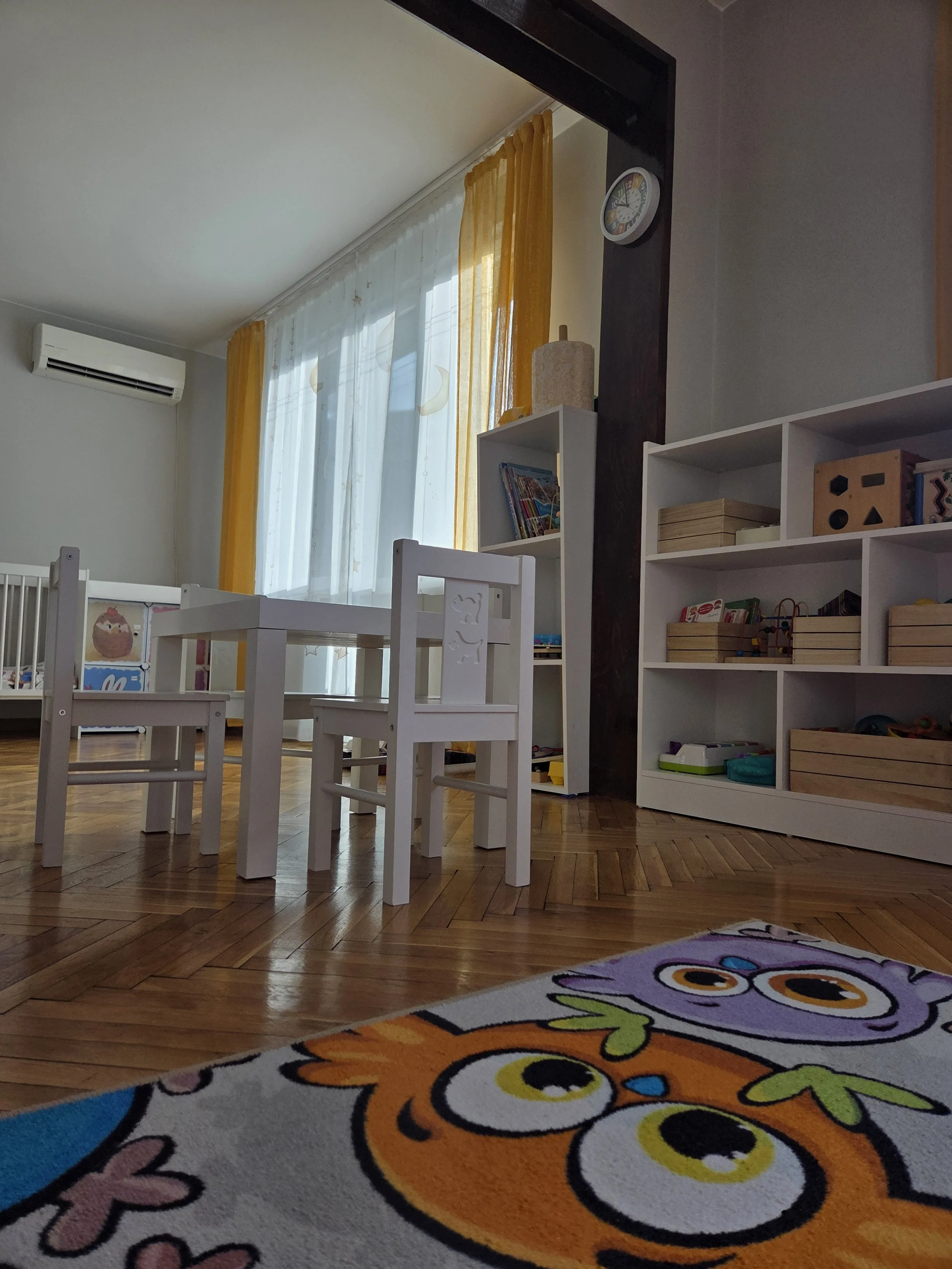 A children's playroom with small white furniture, a bookshelf with toys and books, colorful curtains, and a carpet with cartoon owls.