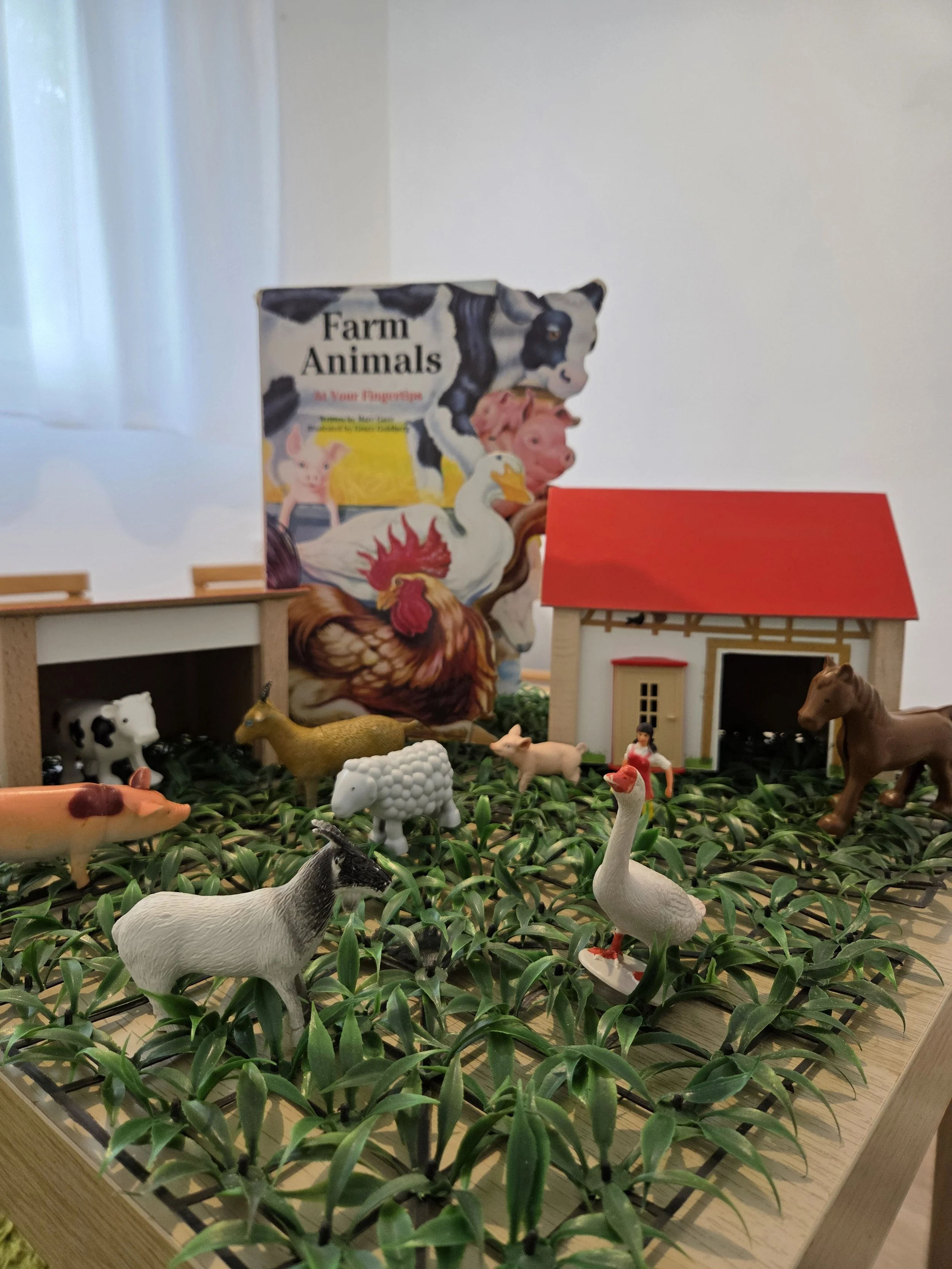 Toy farm scene with plastic animals, a barn, a house, a sunflower book, and green foliage on a wooden table.
