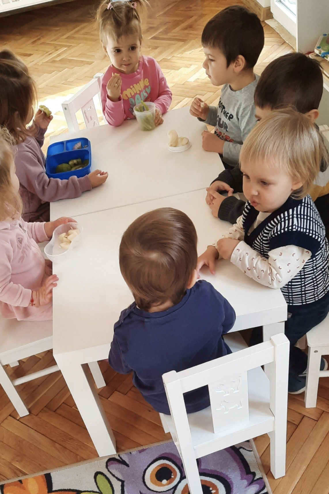 Group of young children sitting around a white table, eating snacks and drinking from cups, in a room with hardwood floors and a partially visible colorful rug.