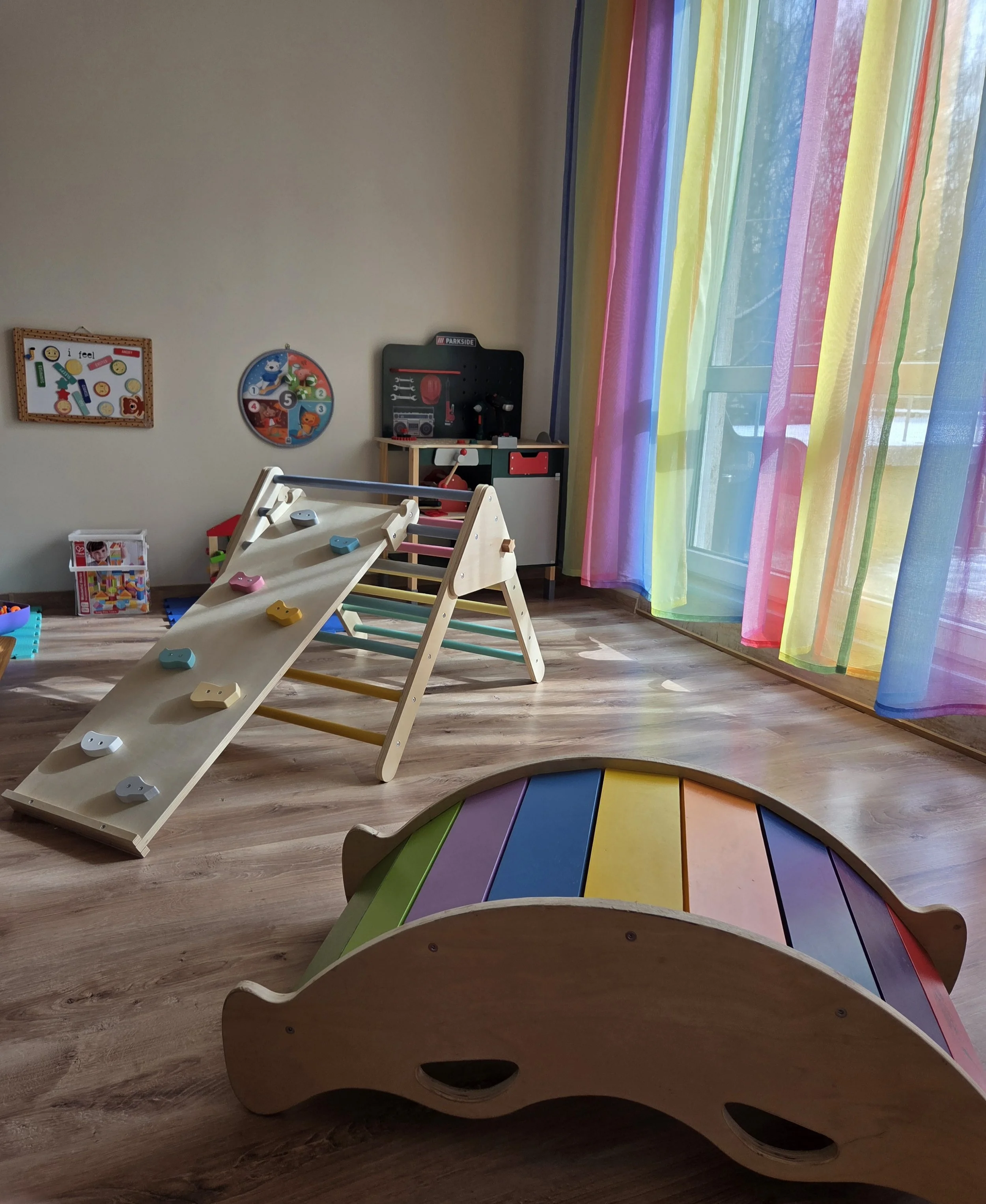 Colorful children's playroom with rainbow curtains, wooden rocking animal with rainbow-colored planks, climbing structure, and various toys.