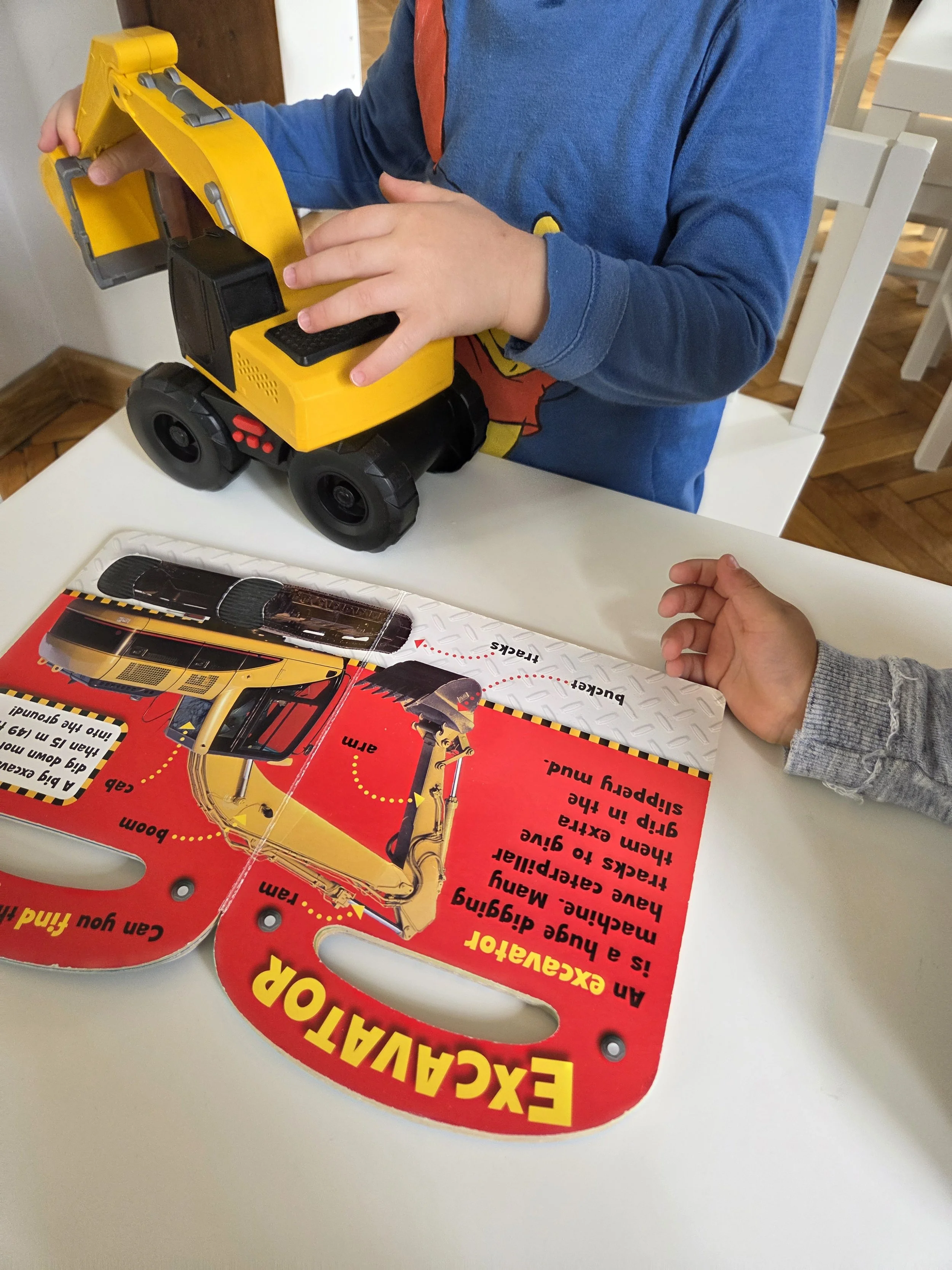 A child playing with a yellow toy excavator on a white table, with an instruction sheet nearby.