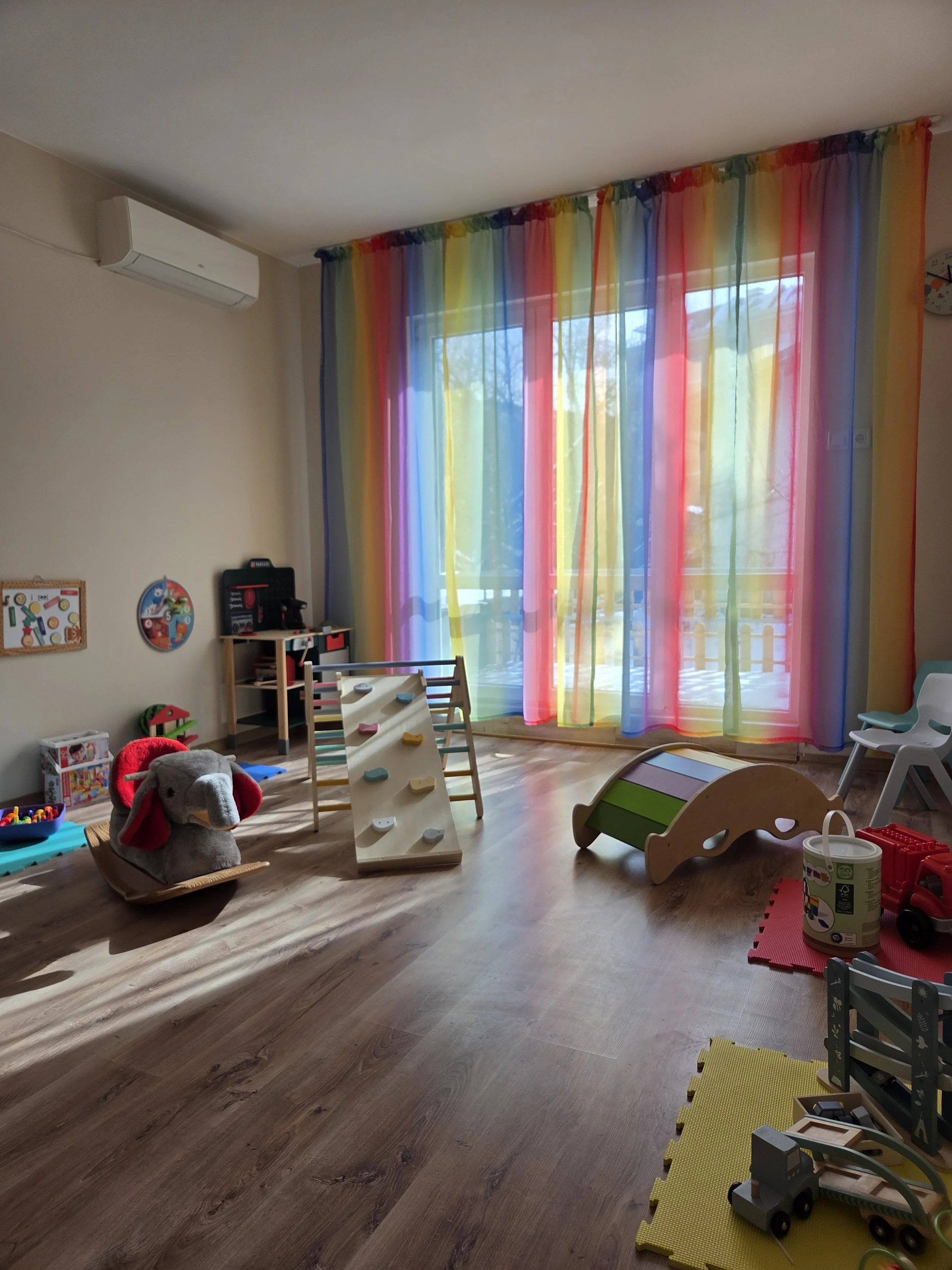 Colorful sheer curtains in rainbow colors covering large window in a child's playroom with toys, a plush elephant, and wooden play structures on a wooden floor.