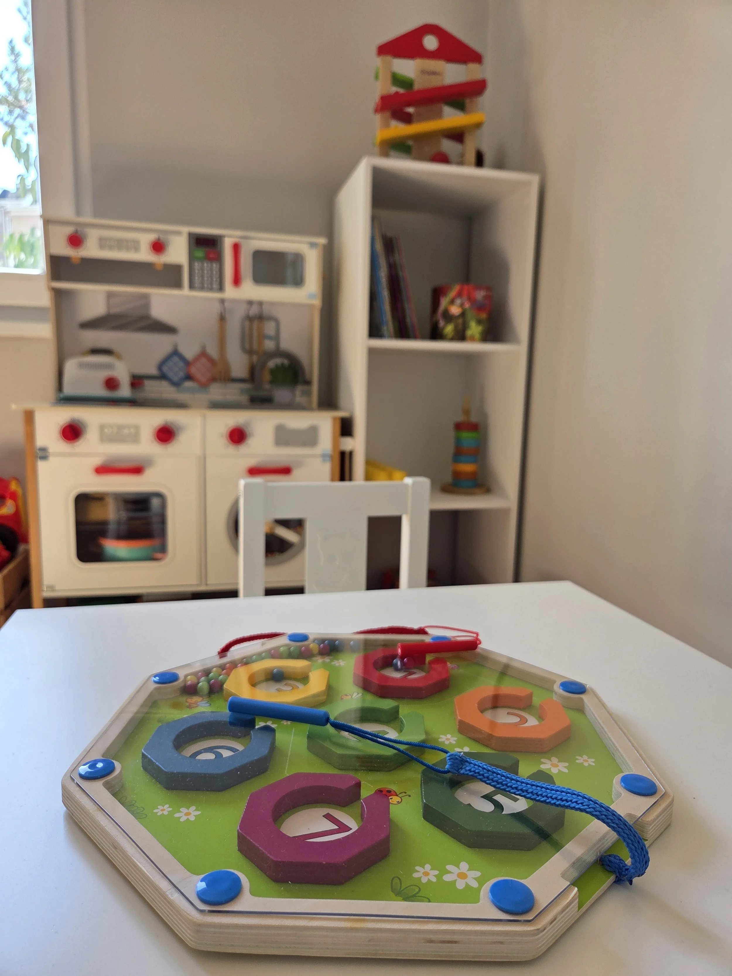 Colorful wooden lacing toy on a white table in a playroom with toy kitchen and bookshelves in the background.