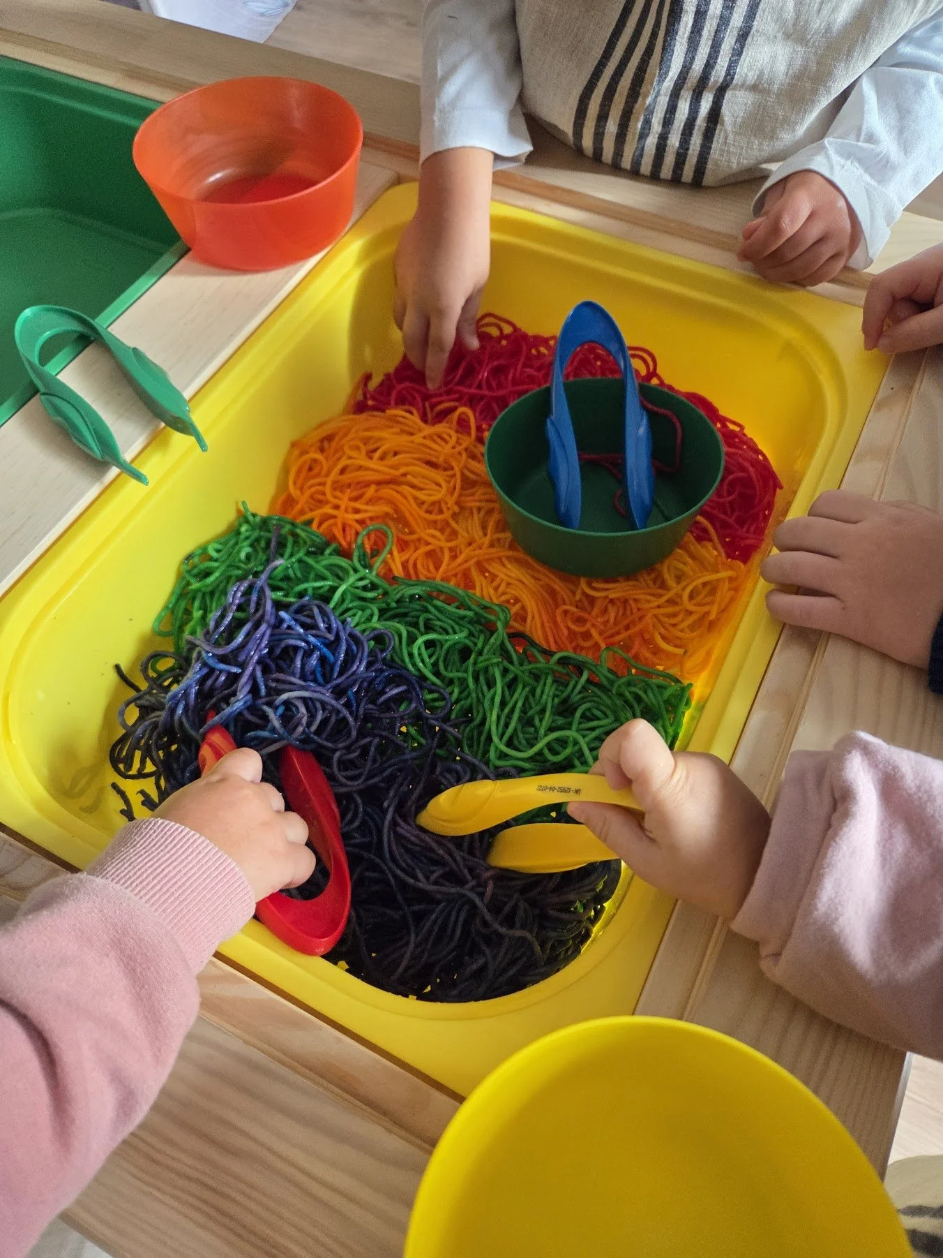 Children playing with colorful plastic scissors and noodle-like material in a sensory bin.