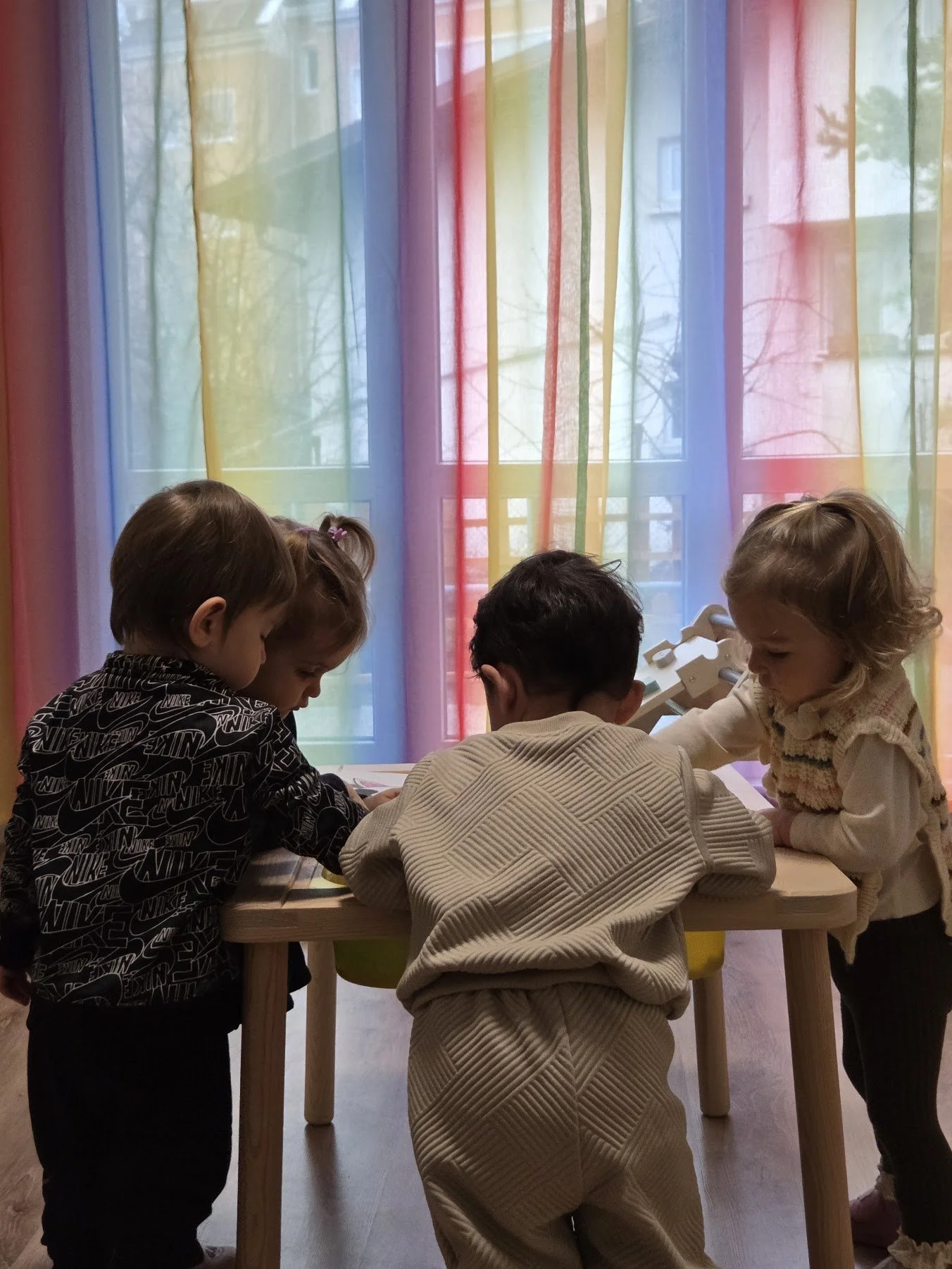 Four children gathered around a table, looking at something together, with a colorful rainbow curtain in the background.