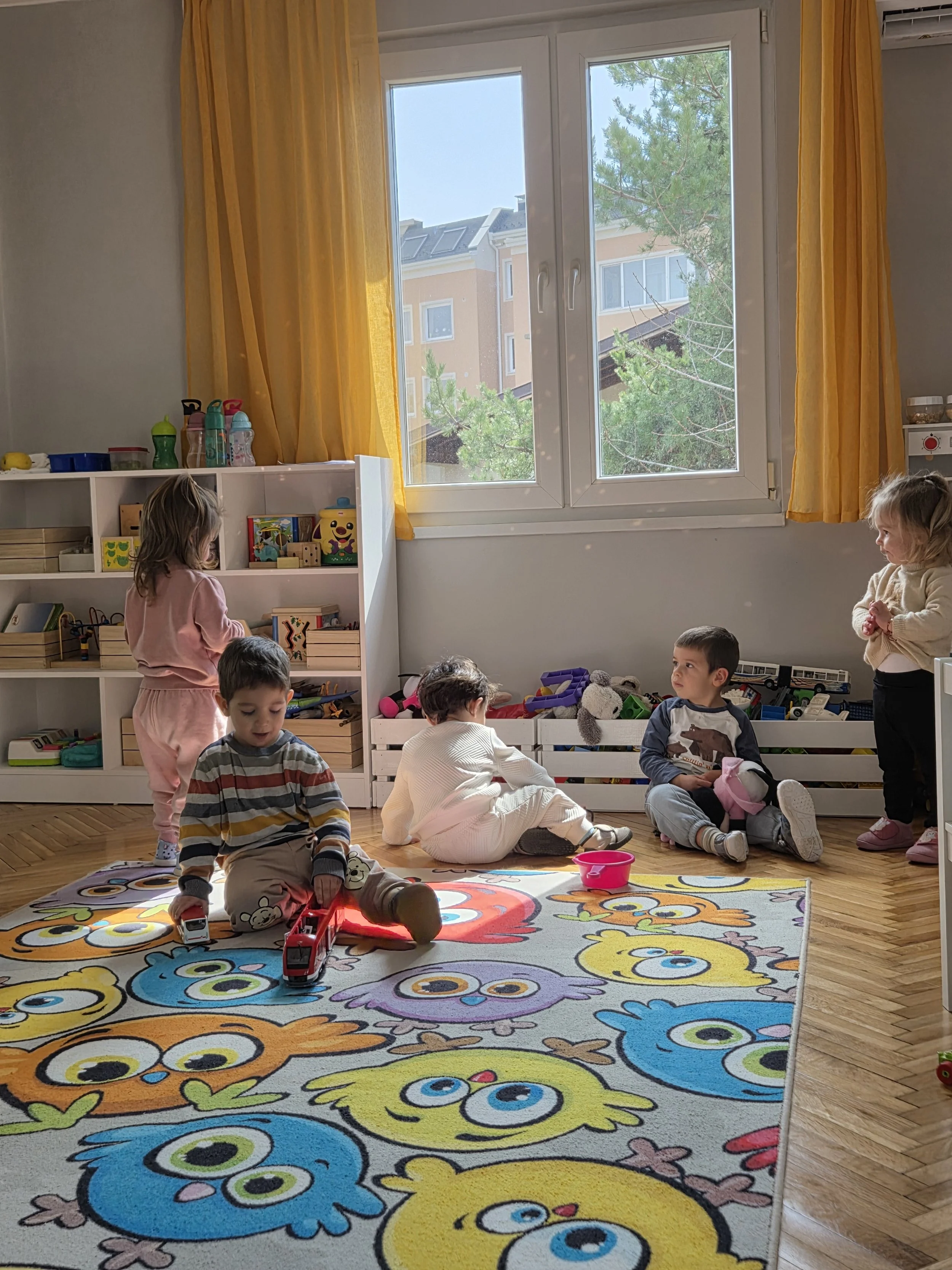 Children playing in a brightly lit room with a colorful owl-patterned rug, toys on a shelf, and a large window with yellow curtains showing trees outside.